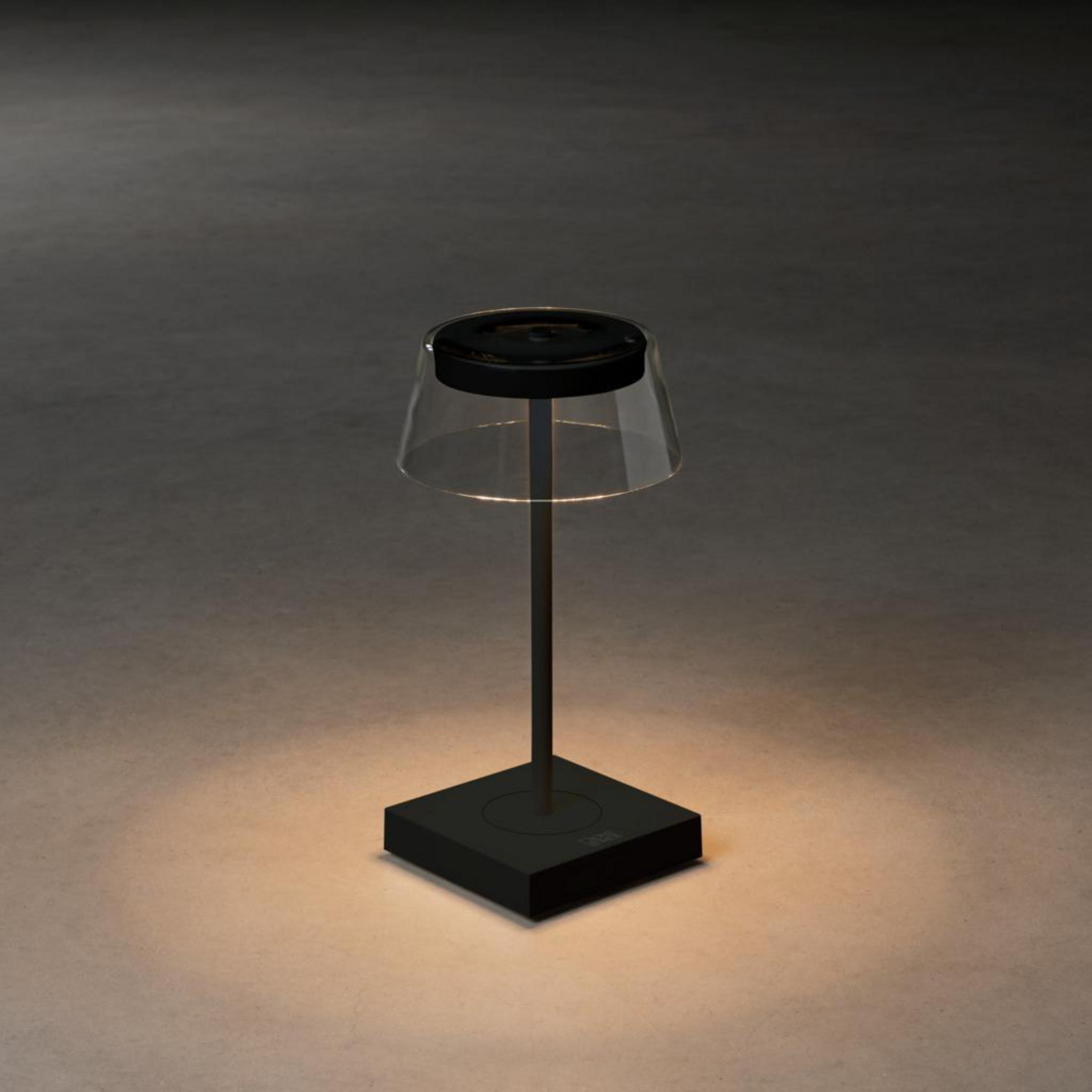 Una lampada da tavolo nera con paralume trasparente e base quadrata, su superficie grigio chiaro.