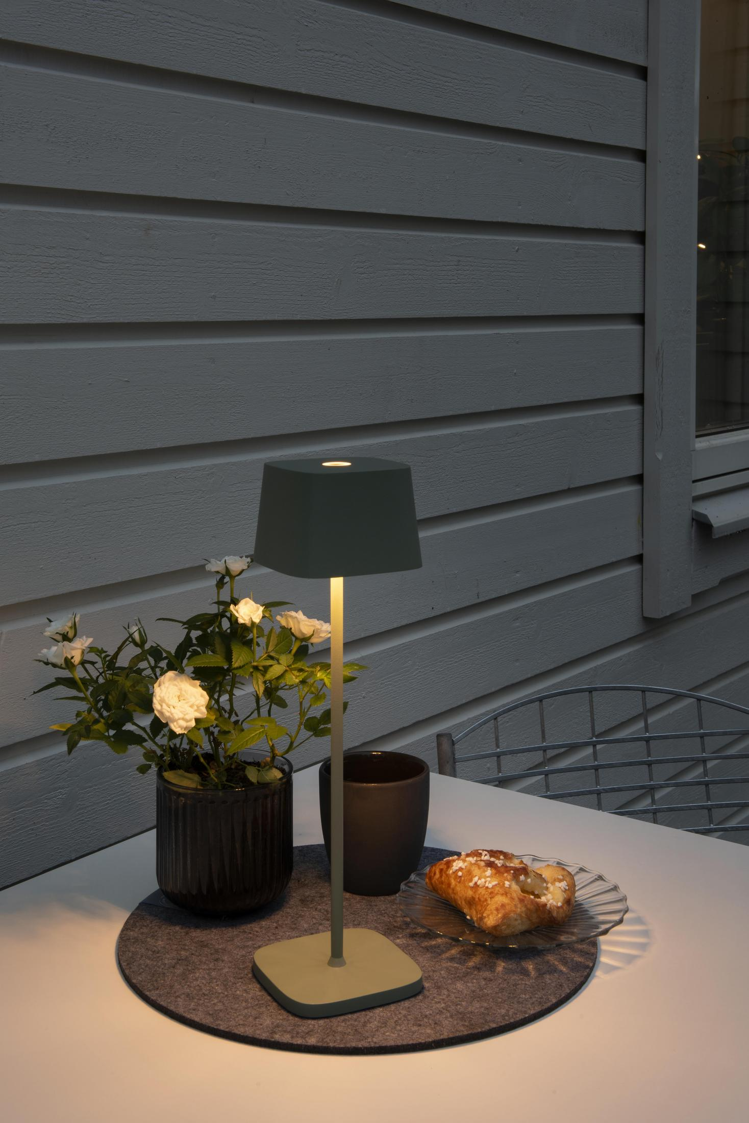 Eine beleuchtete grüne Außenlampe auf einem Tisch, mit Vase, weißen Blumen, einer Tasse und einem Gebäck.