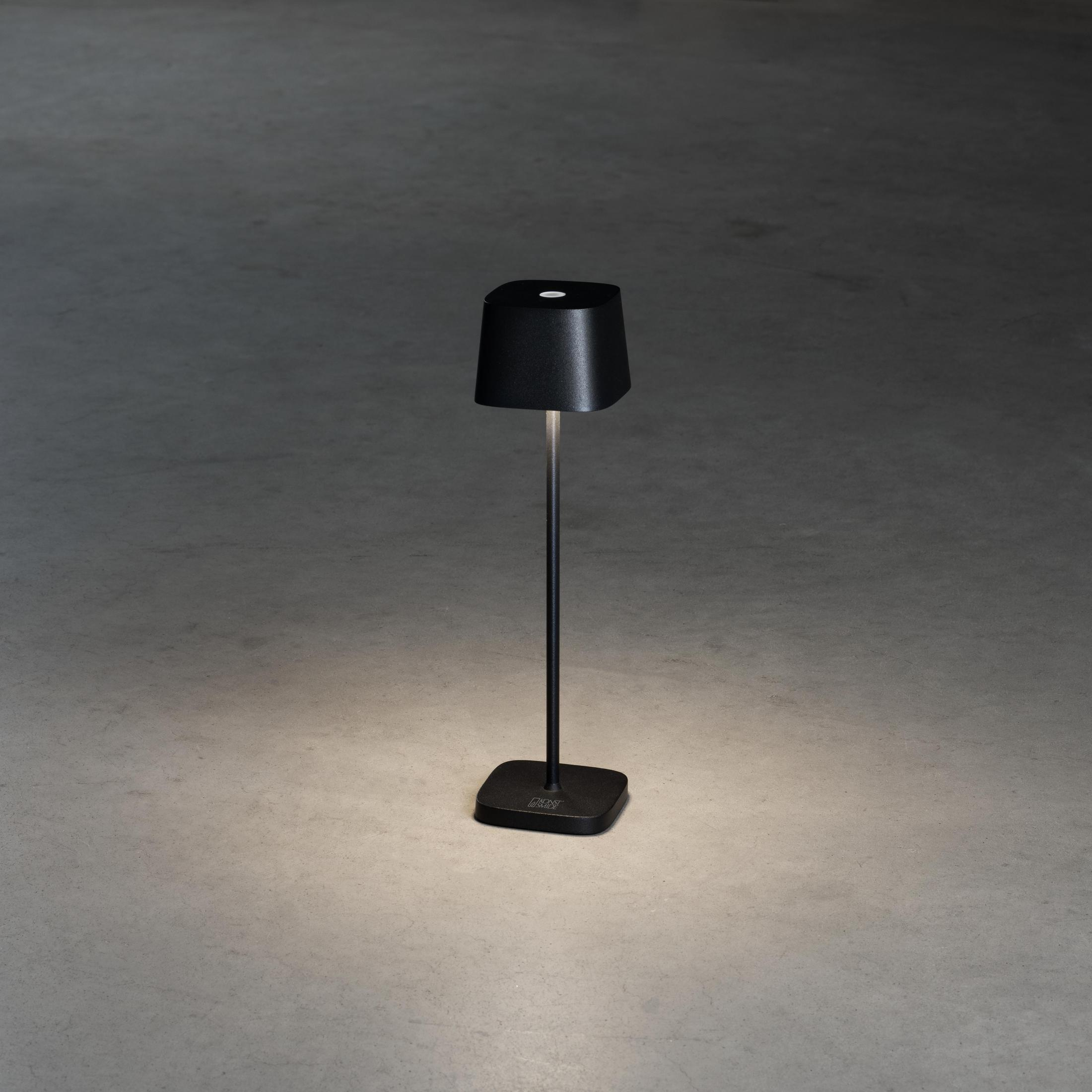 Lampada da tavolo nera, accesa. Luce sul pavimento in cemento. Base nera.