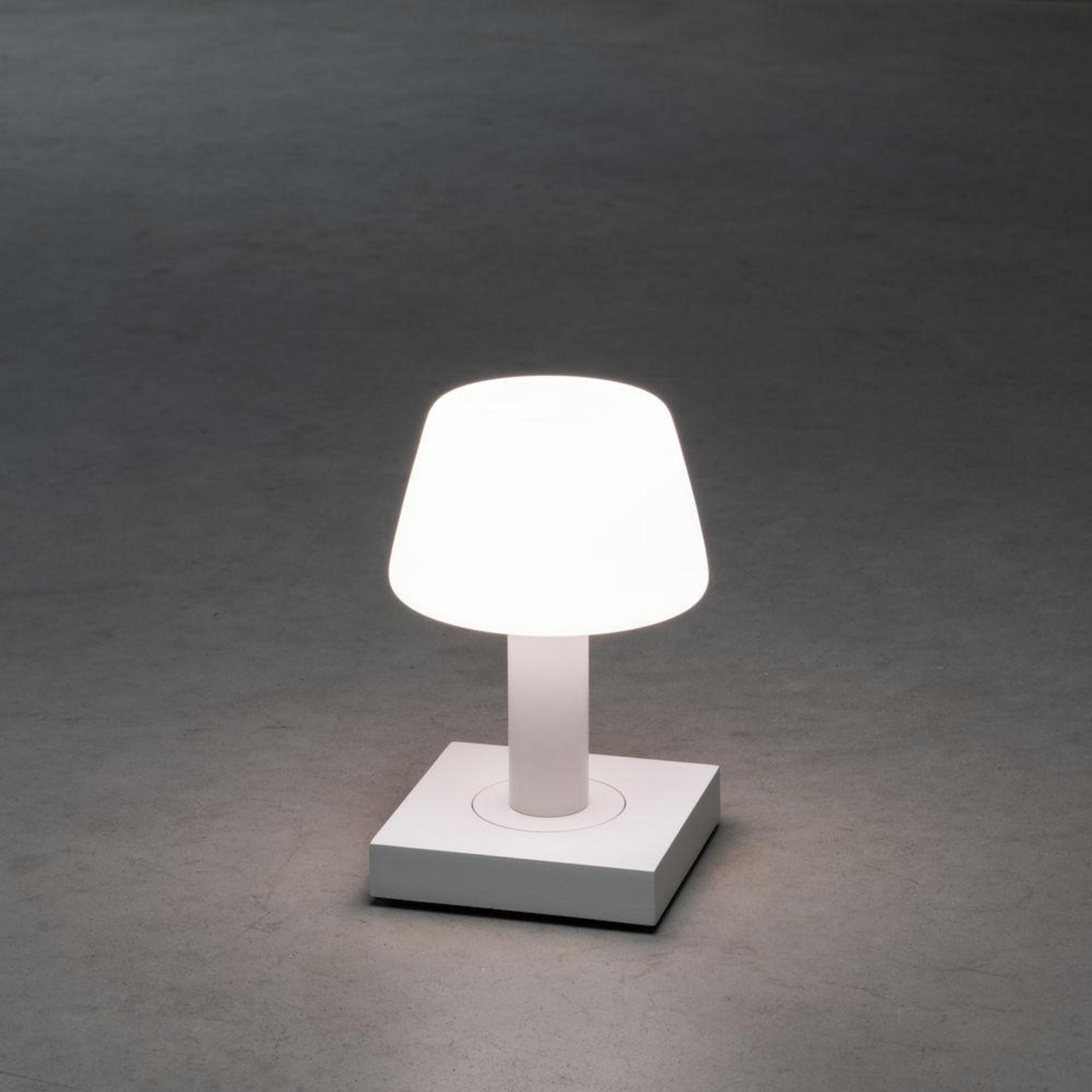 Weiße Lampe mit rundem Oberteil auf weißem Sockel, auf grauem Boden.