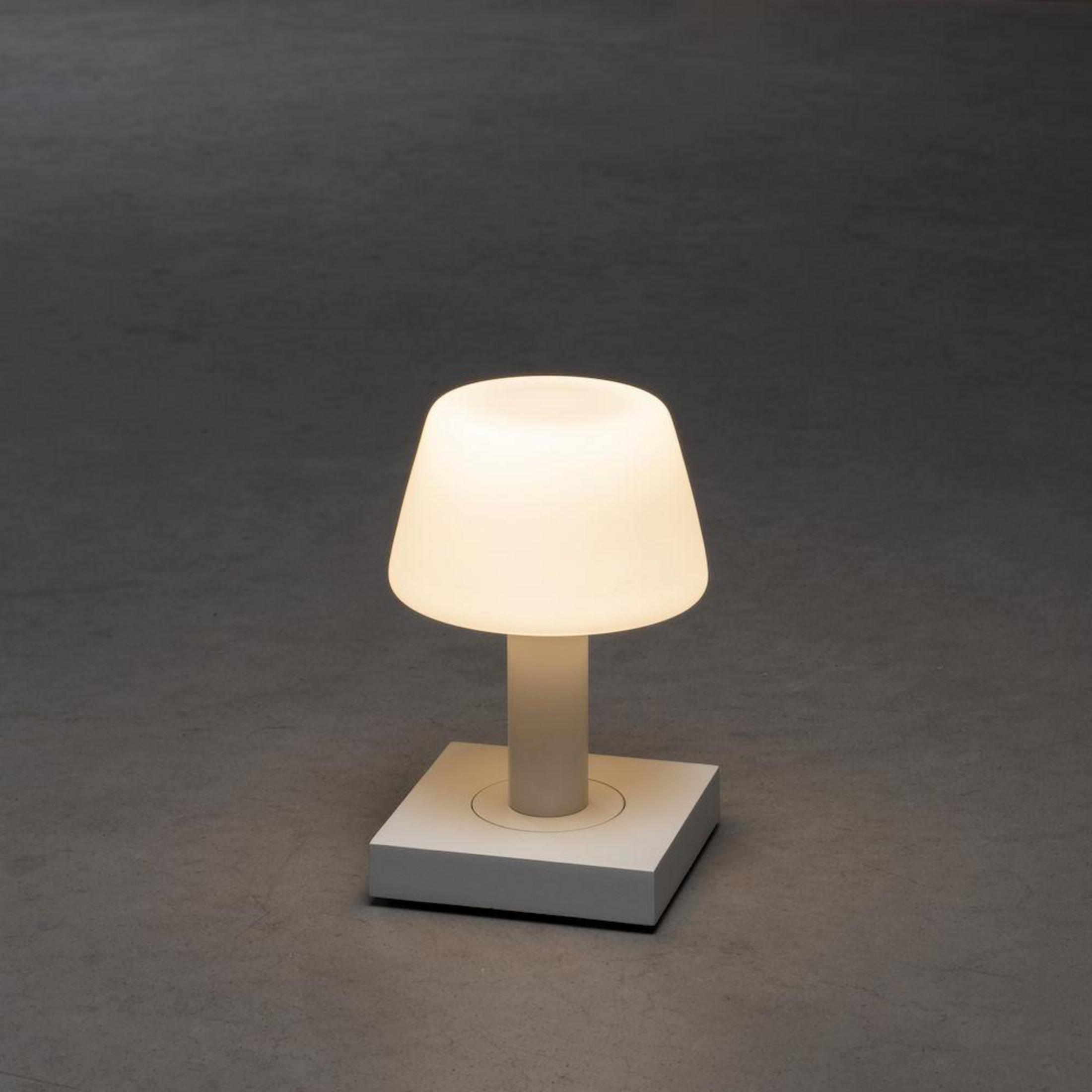 Eine weiße Tischlampe mit Lampenschirm. Die Lampe ist eingeschaltet und strahlt helles Licht aus. Sie steht auf grauem Hintergrund.