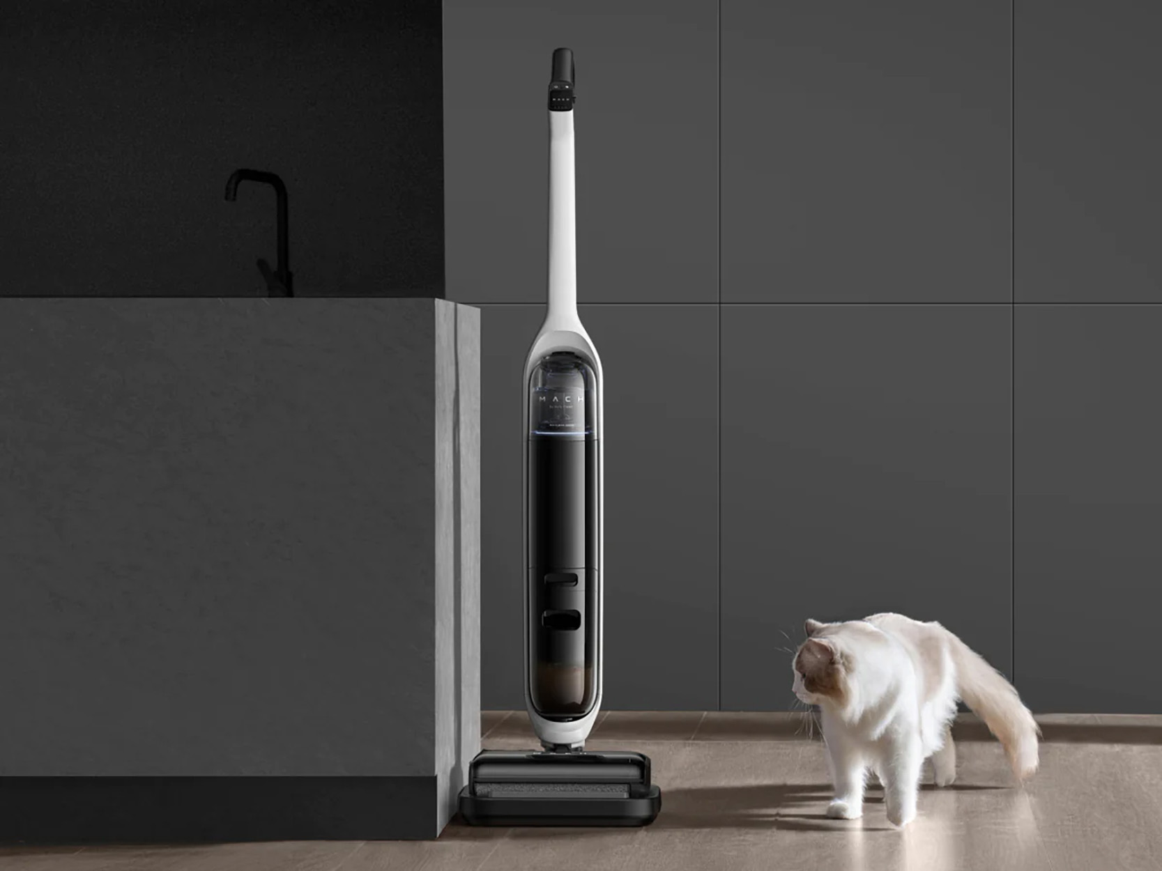 Een witte en zwarte stofzuiger staat naast een kat in een grijze kamer.