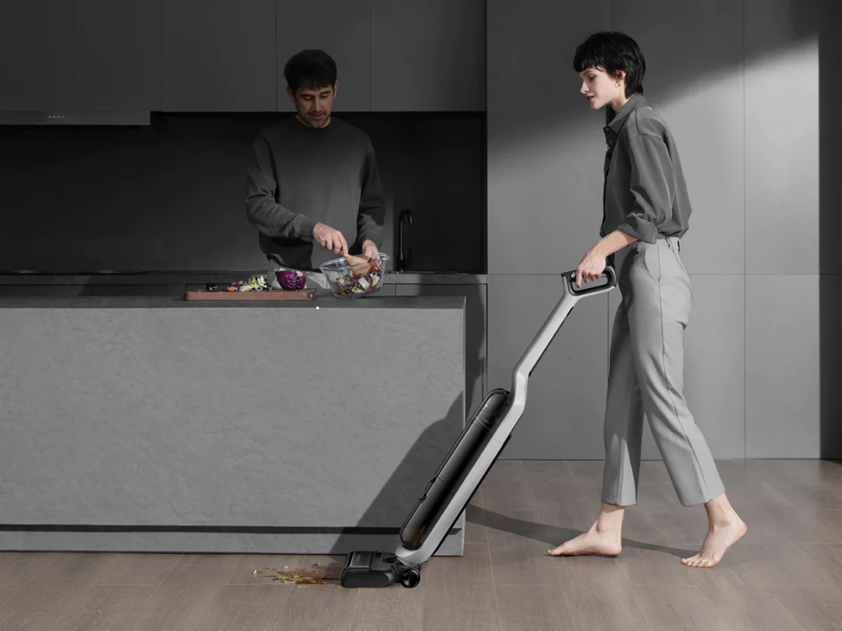 Een vrouw stofzuigt de vloer, terwijl een man eten bereidt in een moderne keuken. Houten vloer.