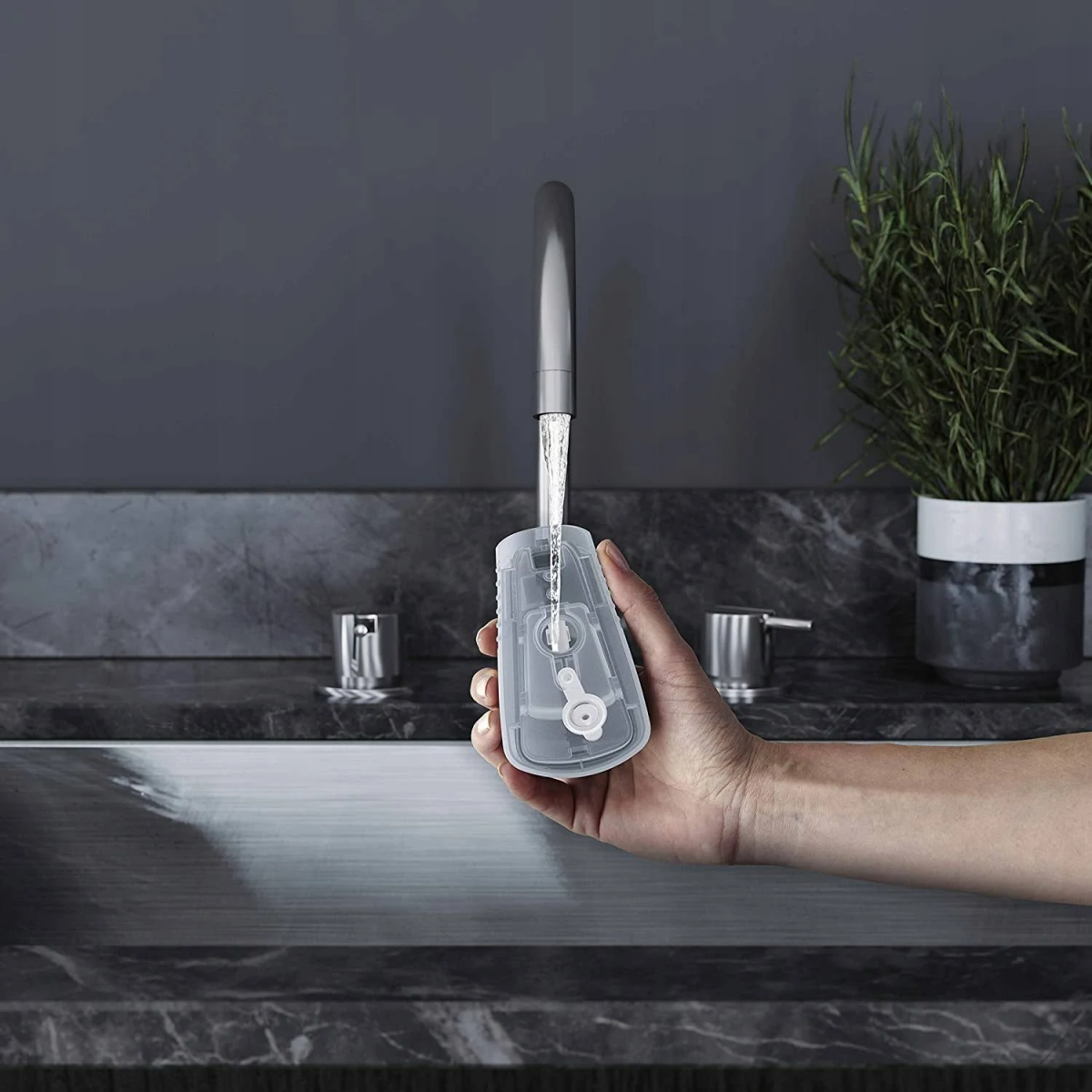 Mano che tiene un dispositivo di plastica trasparente con acqua che scorre. Rubinetto e piante sullo sfondo.