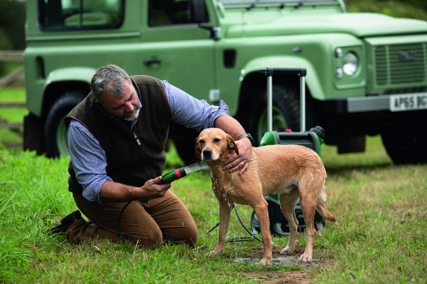 Ein Mann wäscht einen Hund mit einem Schlauch auf einer Wiese, ein grüner SUV im Hintergrund.
