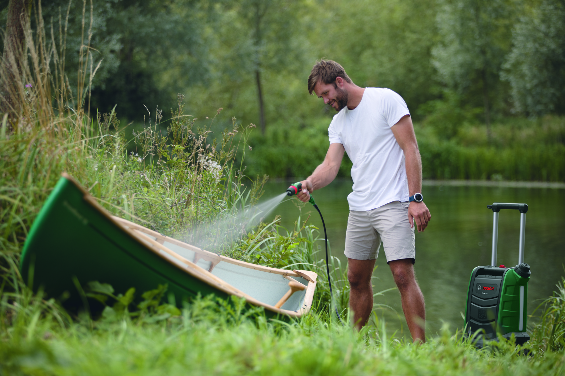 Ein Mann wäscht ein grünes Kanu im Wasser, mit einem Hochdruckreiniger daneben.