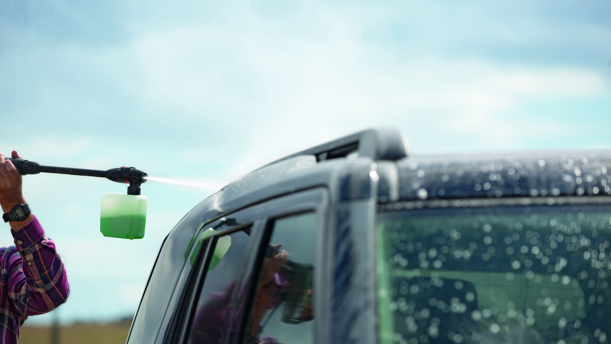 Personne lavant une voiture noire avec un nettoyeur haute pression. Mousse verte et ciel bleu.