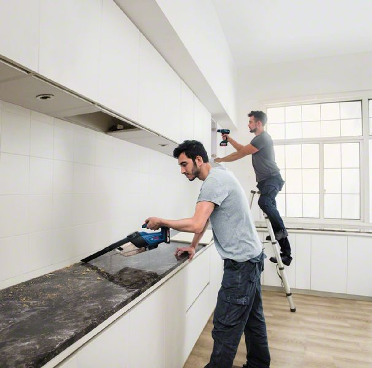 Dwóch mężczyzn w kuchni, jeden używa odkurzacza, drugi na drabinie używa wiertarki.