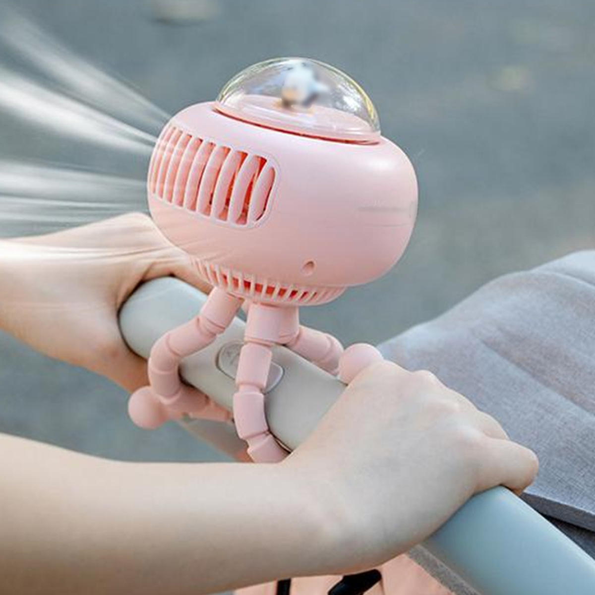 Hände einer Person halten einen rosa Ventilator mit einer durchsichtigen Kuppel, der an einem Kinderwagen befestigt ist.