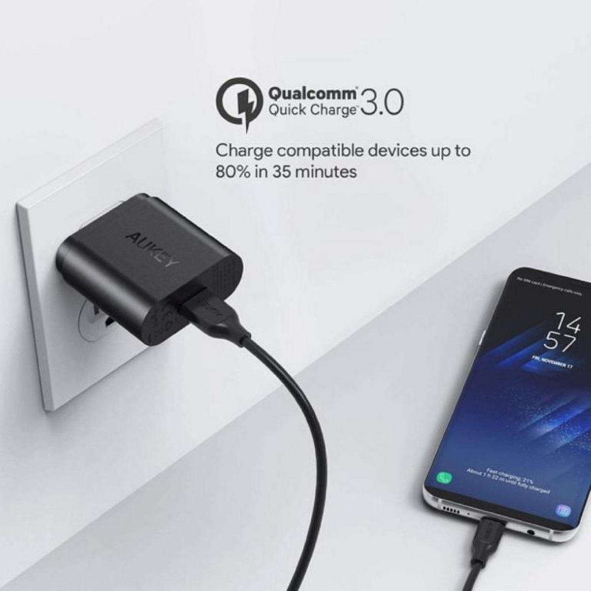 Schwarzes AUKEY-Ladegerät in einer weißen Steckdose. Ein Telefon wird in der Nähe geladen.