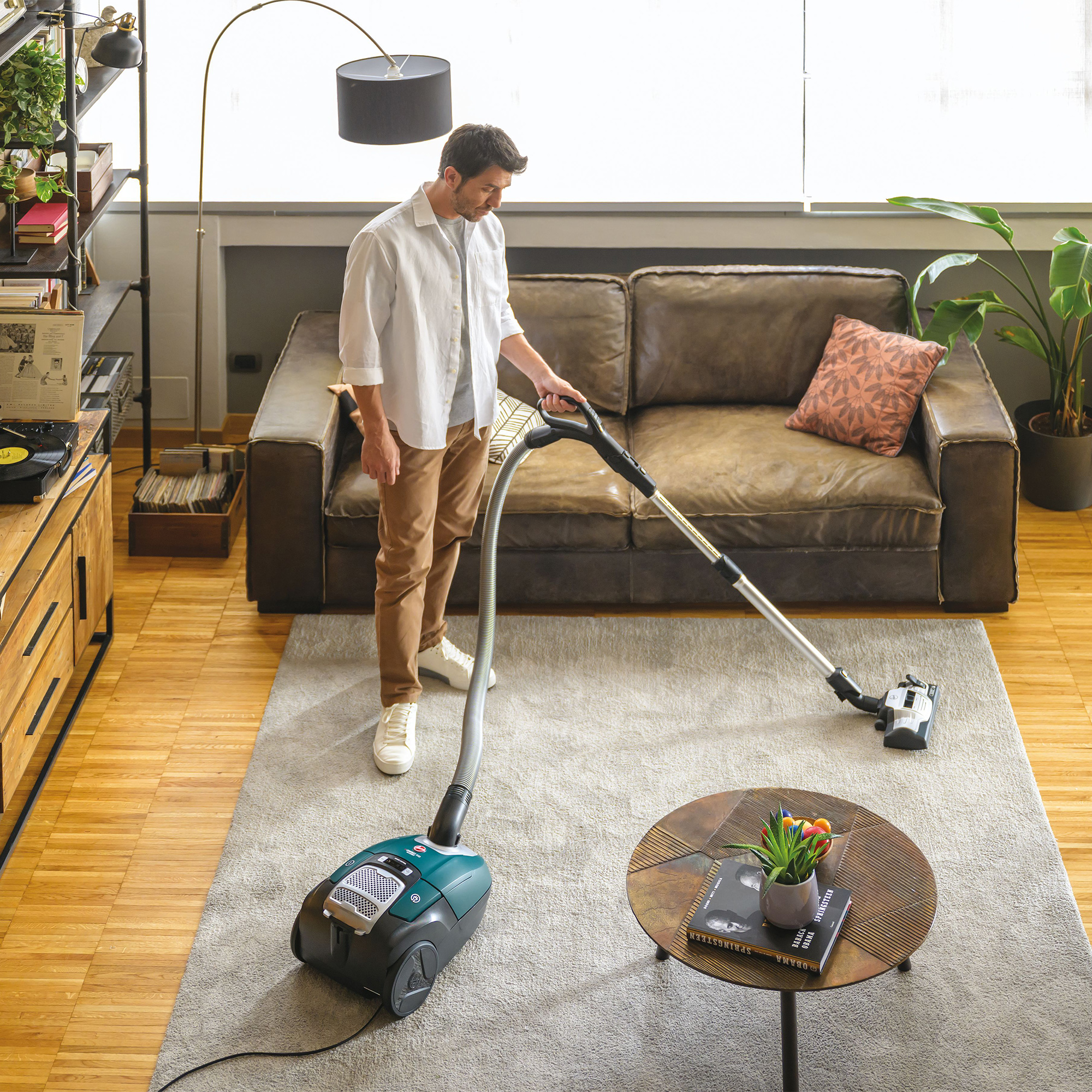 Mężczyzna odkurza dywan w salonie. Brązowa sofa, stolik i regał w tle.