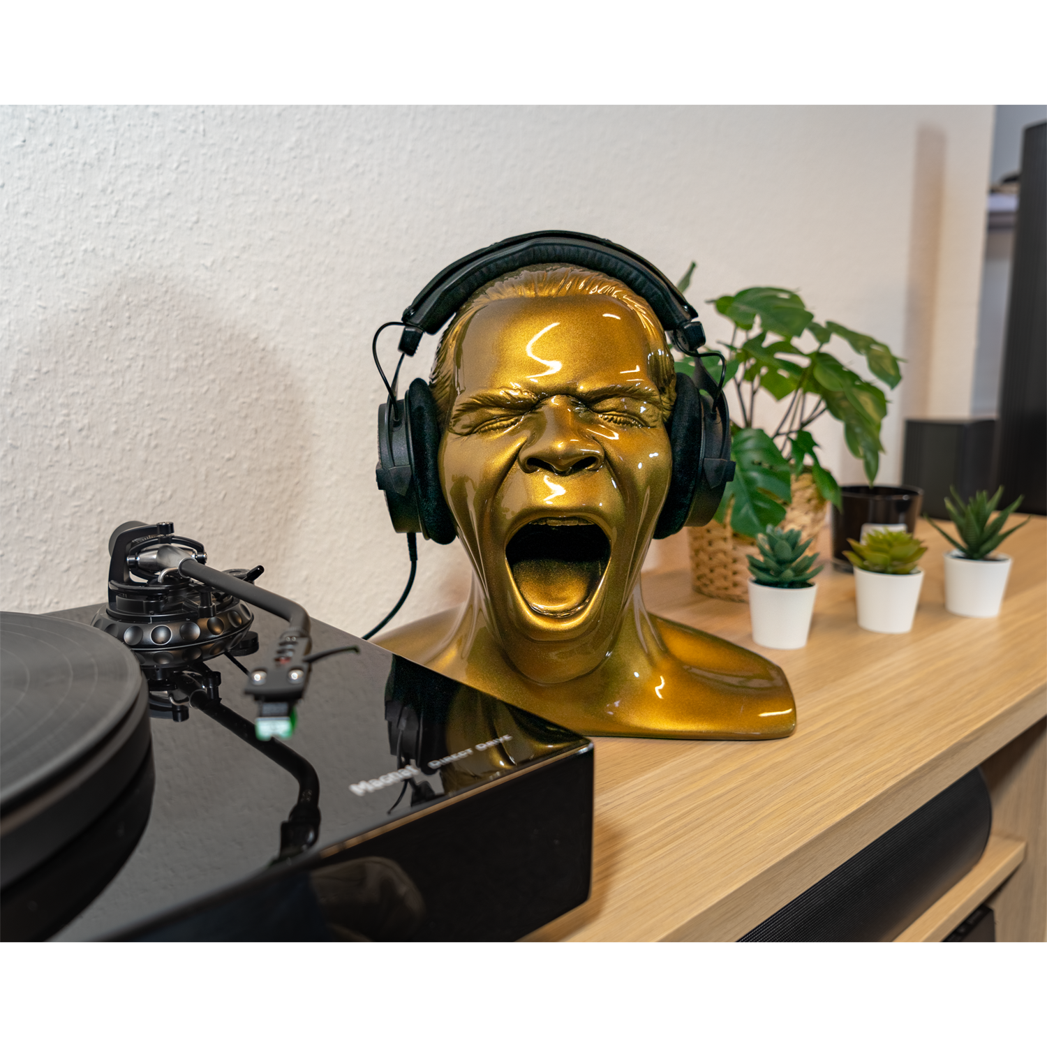 Goldene Kopfskulptur mit Kopfhörern auf einem Holztisch. Ein schwarzer Plattenspieler daneben.