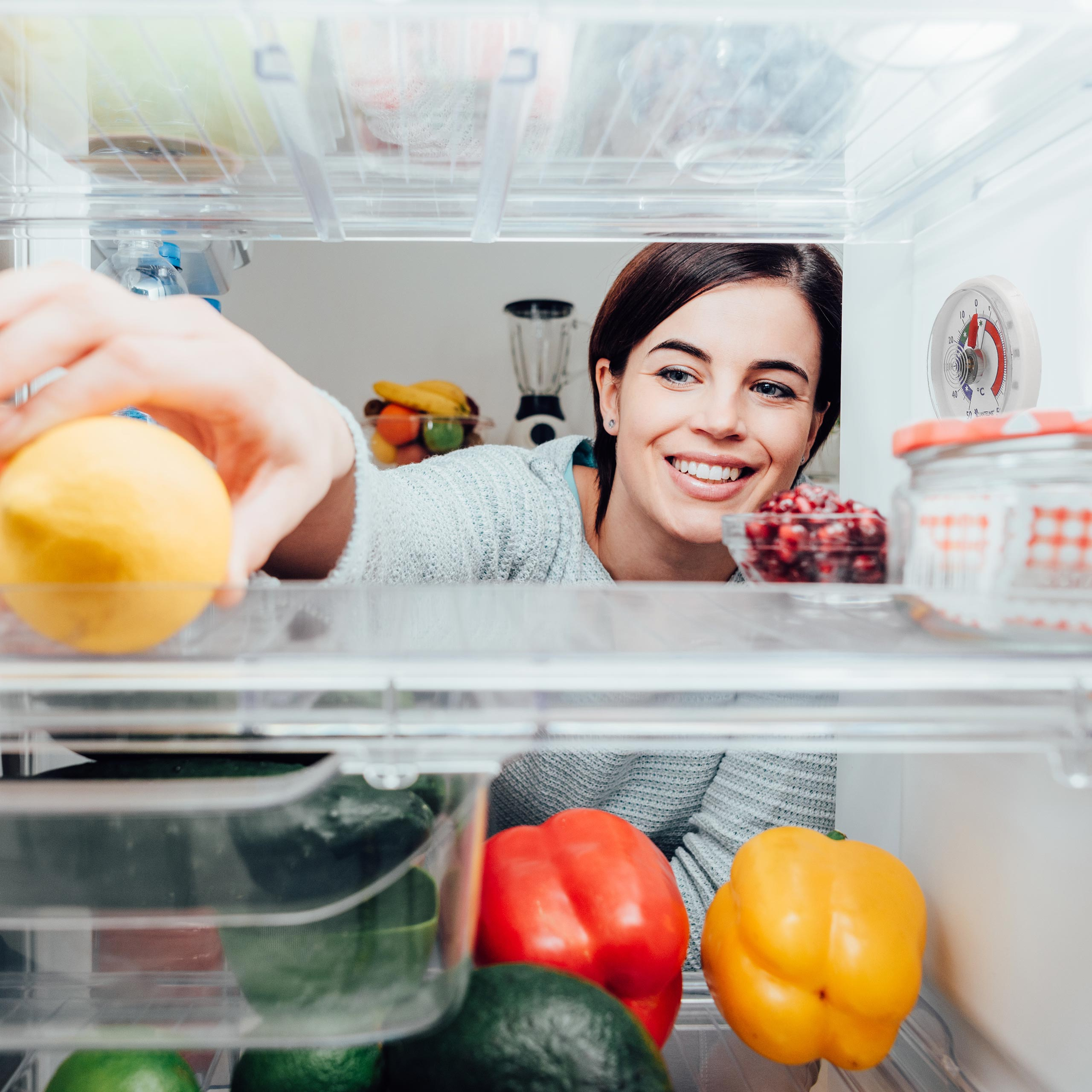 Eine Frau im Kühlschrank, die nach einer Zitrone greift, mit anderen Früchten und Regalen sichtbar.
