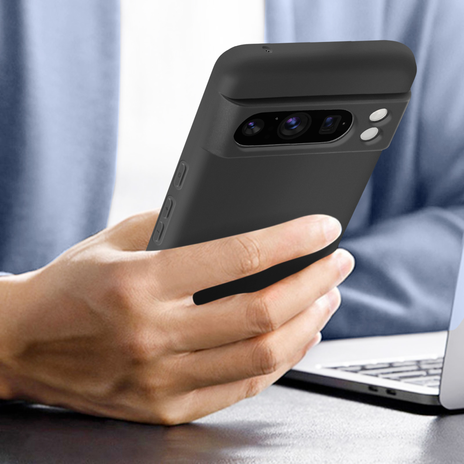 Una mano che tiene un telefono nero in una custodia scura. Un laptop e una persona sfocata sullo sfondo.