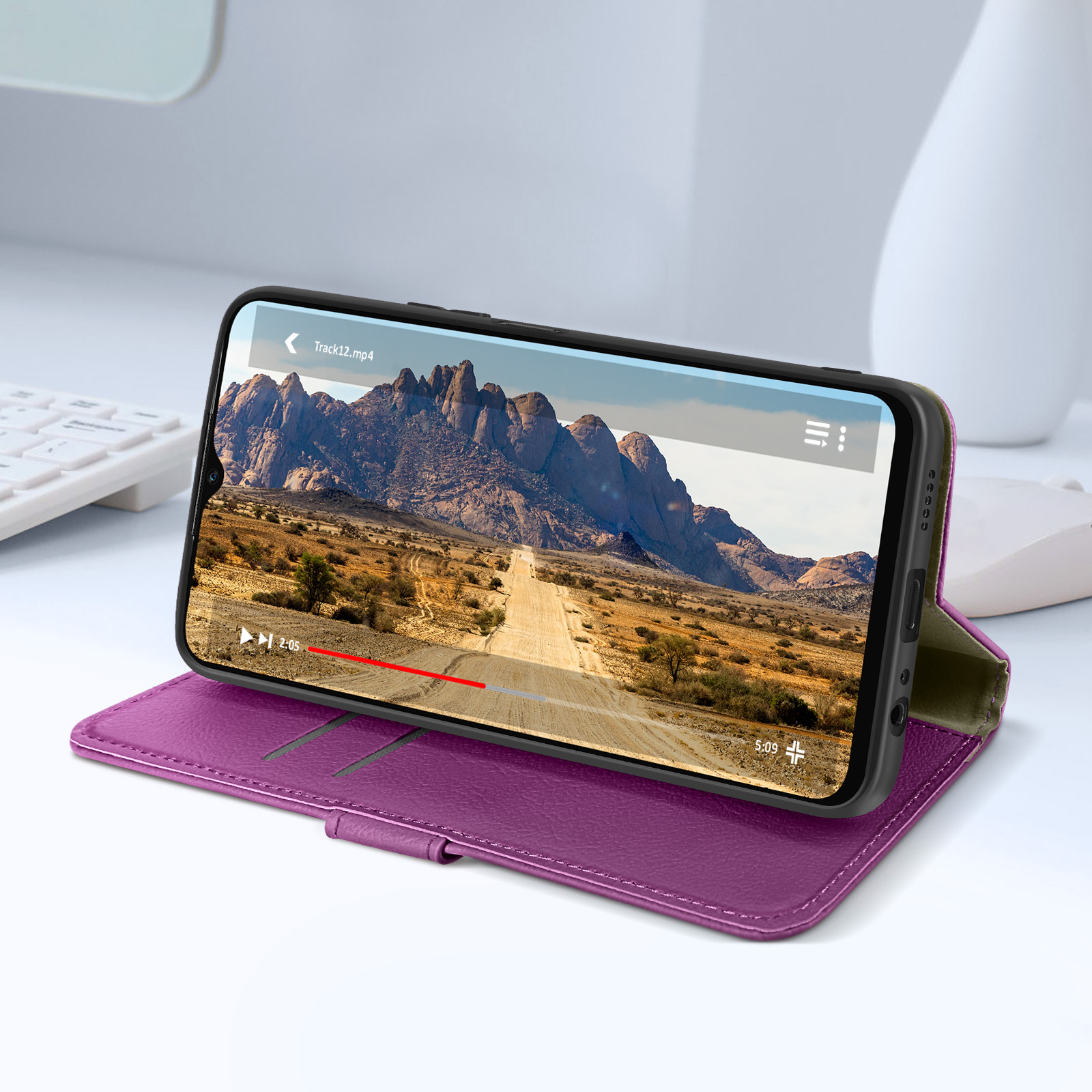 Telefono in custodia viola mostra video di una strada. Scrivania bianca, vaso e tastiera sullo sfondo.