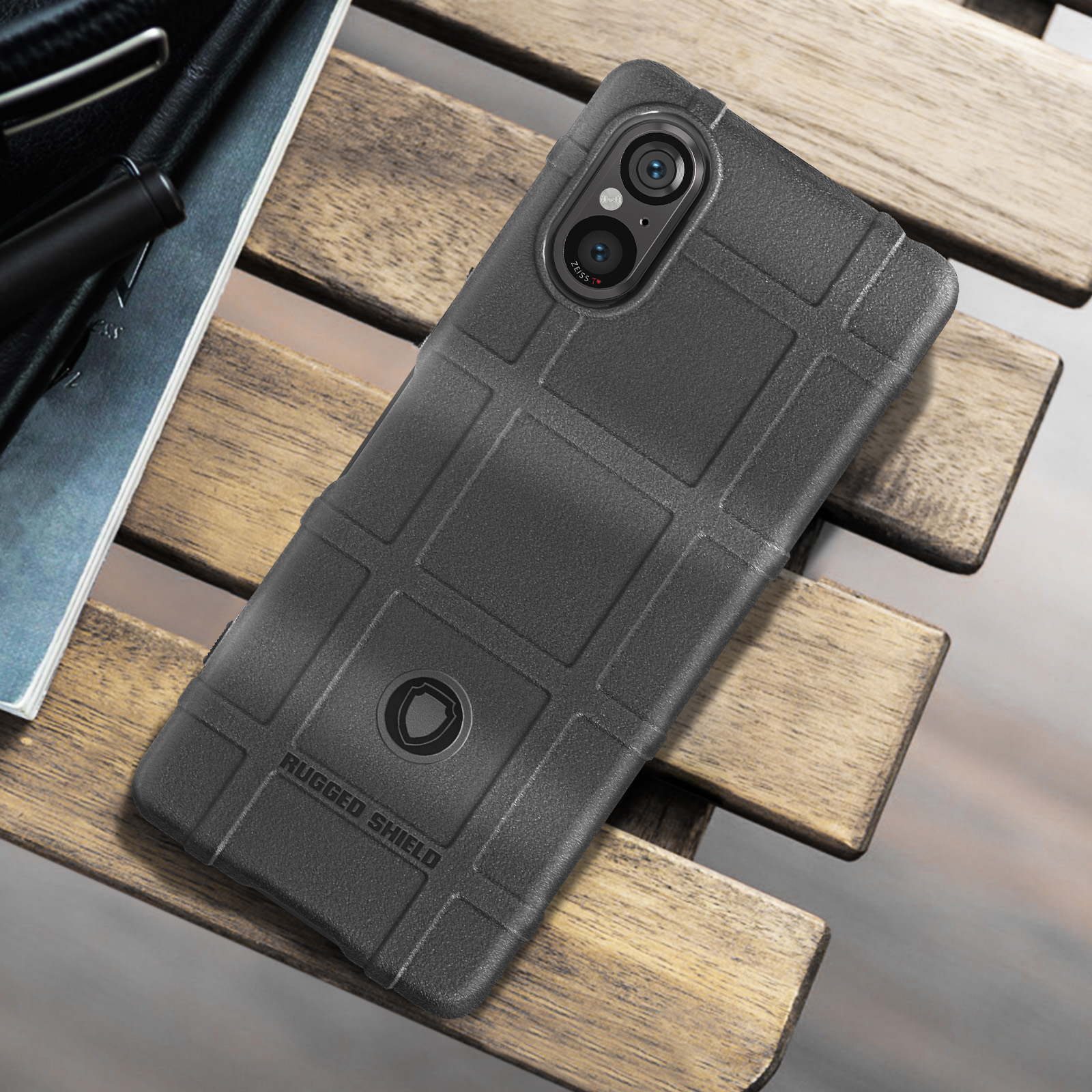 Una custodia nera con motivo a griglia, appoggiata su un tavolo di legno. La custodia ha 'RUGGED SHIELD'.