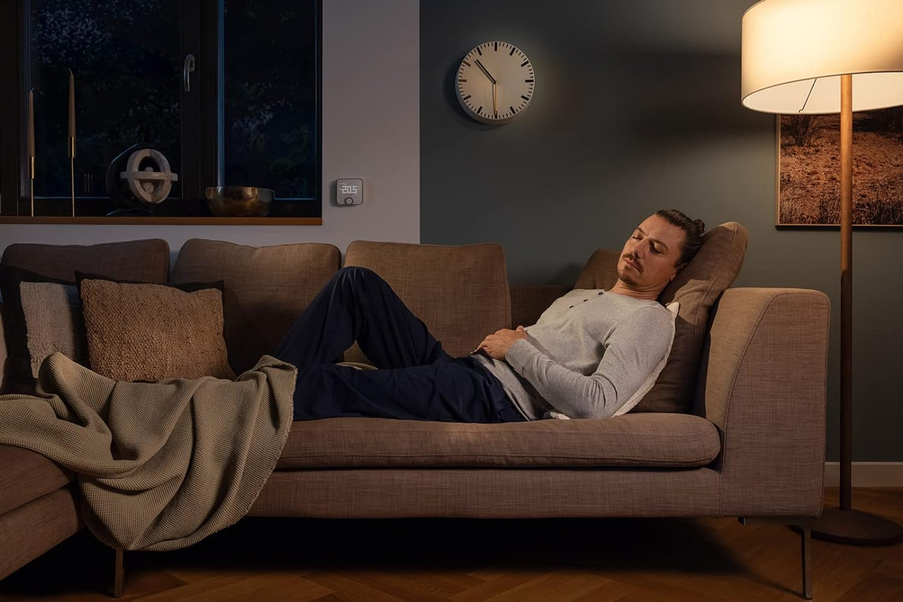 Mann ruht sich auf einer Couch im Wohnzimmer aus. Im Hintergrund befinden sich eine Uhr, eine Lampe und ein Fenster.