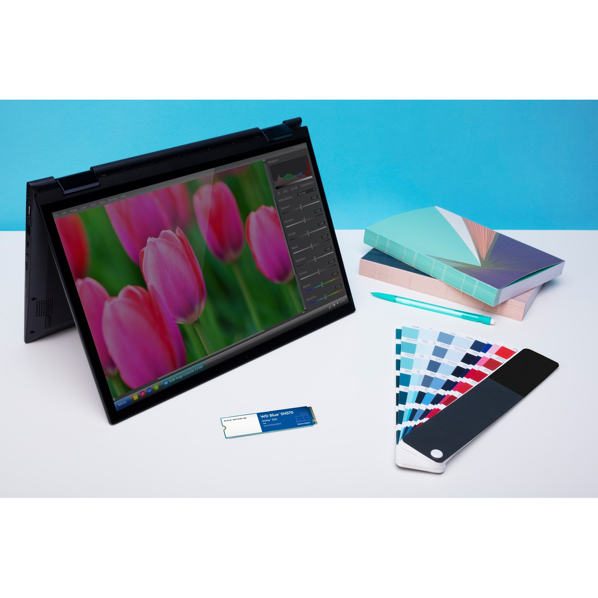 Laptop wyświetlający różowy obraz kwiatowy na niebieskim tle. Niebieski dysk SSD i palety kolorów leżą na stole.