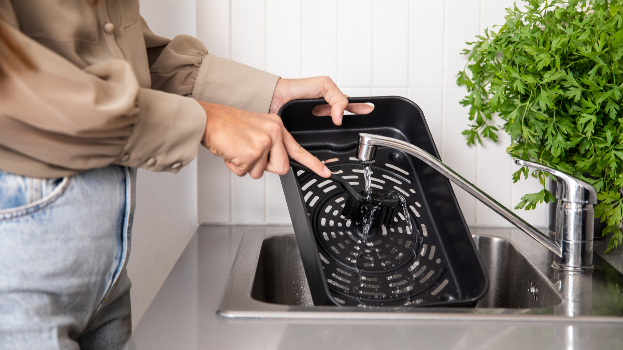 Osoba myje czarny kosz do frytkownicy beztłuszczowej pod bieżącą wodą. Wnętrze jest perforowane.
