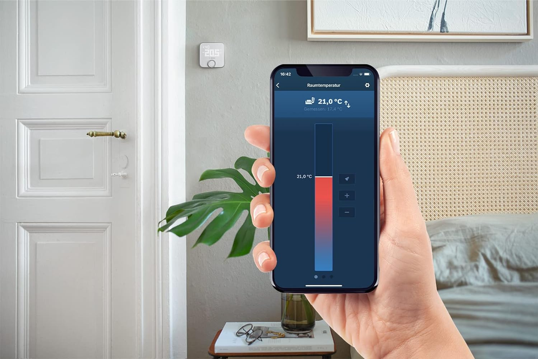 Eine Hand hält ein Smartphone mit geöffneter Temperatur-App. Im Hintergrund eine Tür und ein Bett.