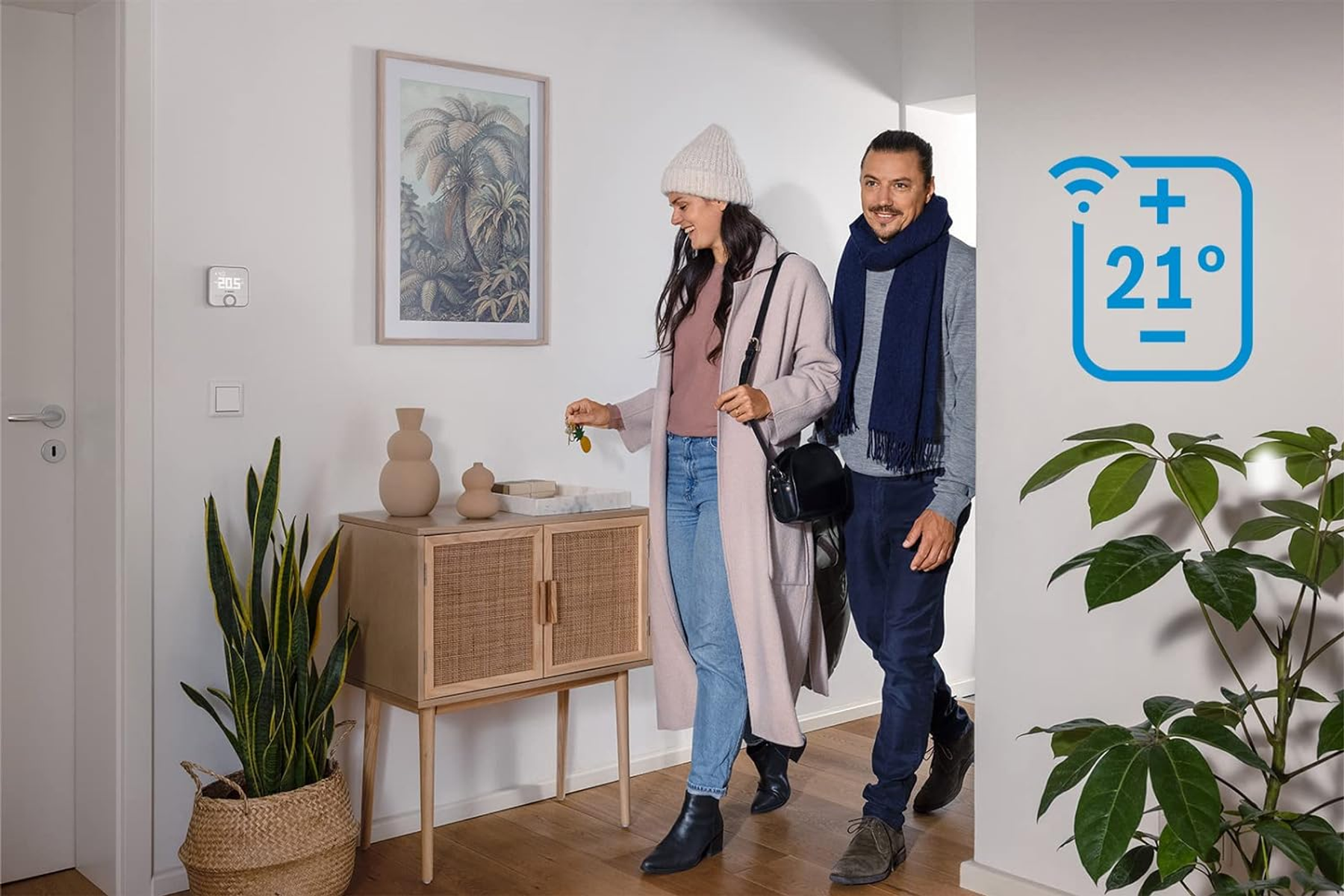 Ein Paar betritt ein Zuhause mit einem intelligenten Thermostat an der Wand. Das Thermostat zeigt 21 Grad an.