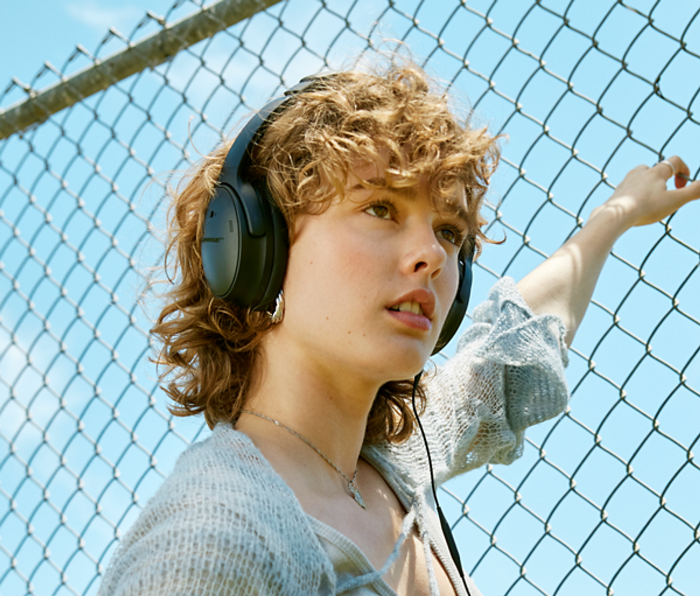 Una mujer con auriculares, de pie junto a una valla de alambre y mirando hacia un lado.