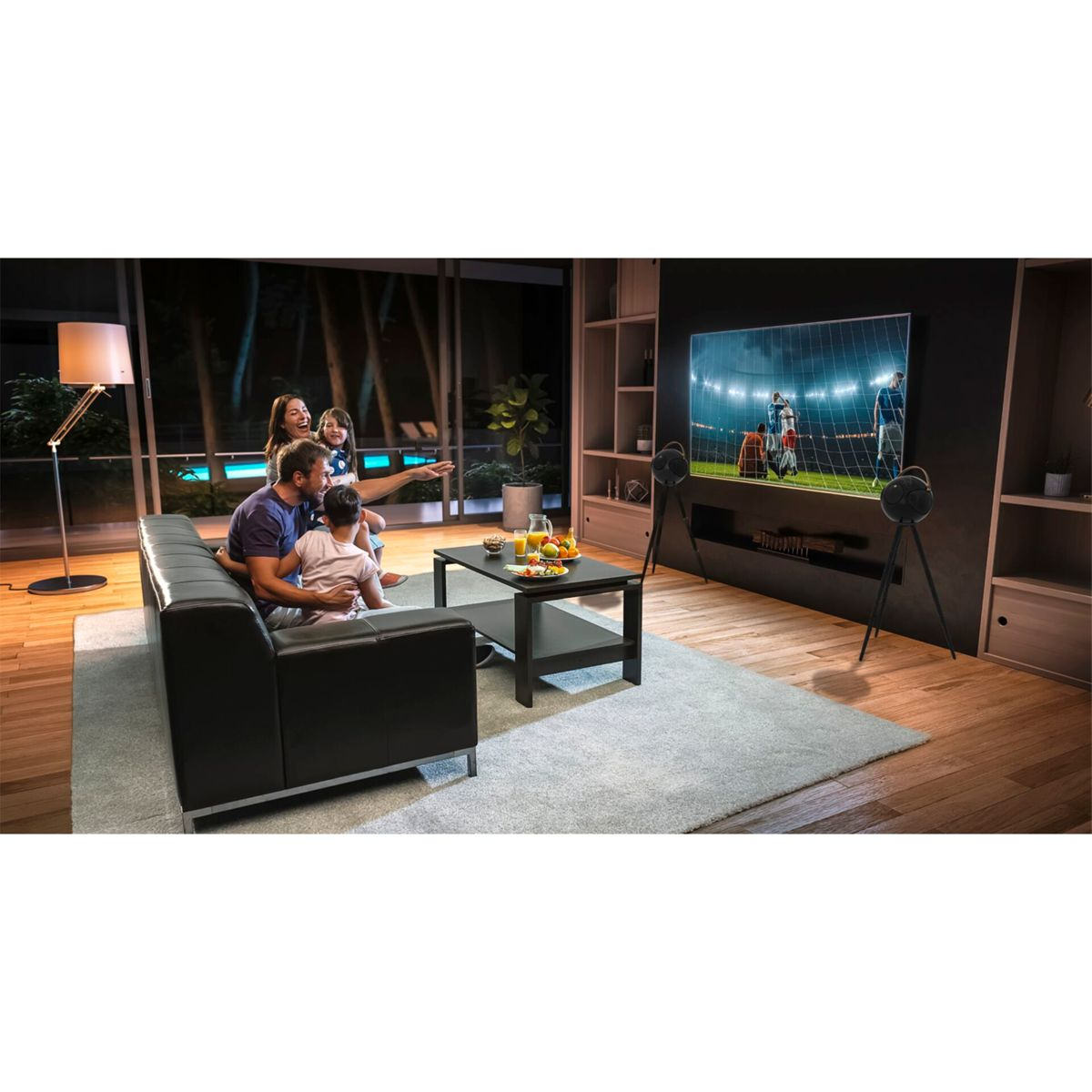 Rodzina ogląda telewizję w salonie. Telewizor pokazuje mecz piłki nożnej. Jest lampa i stolik kawowy.