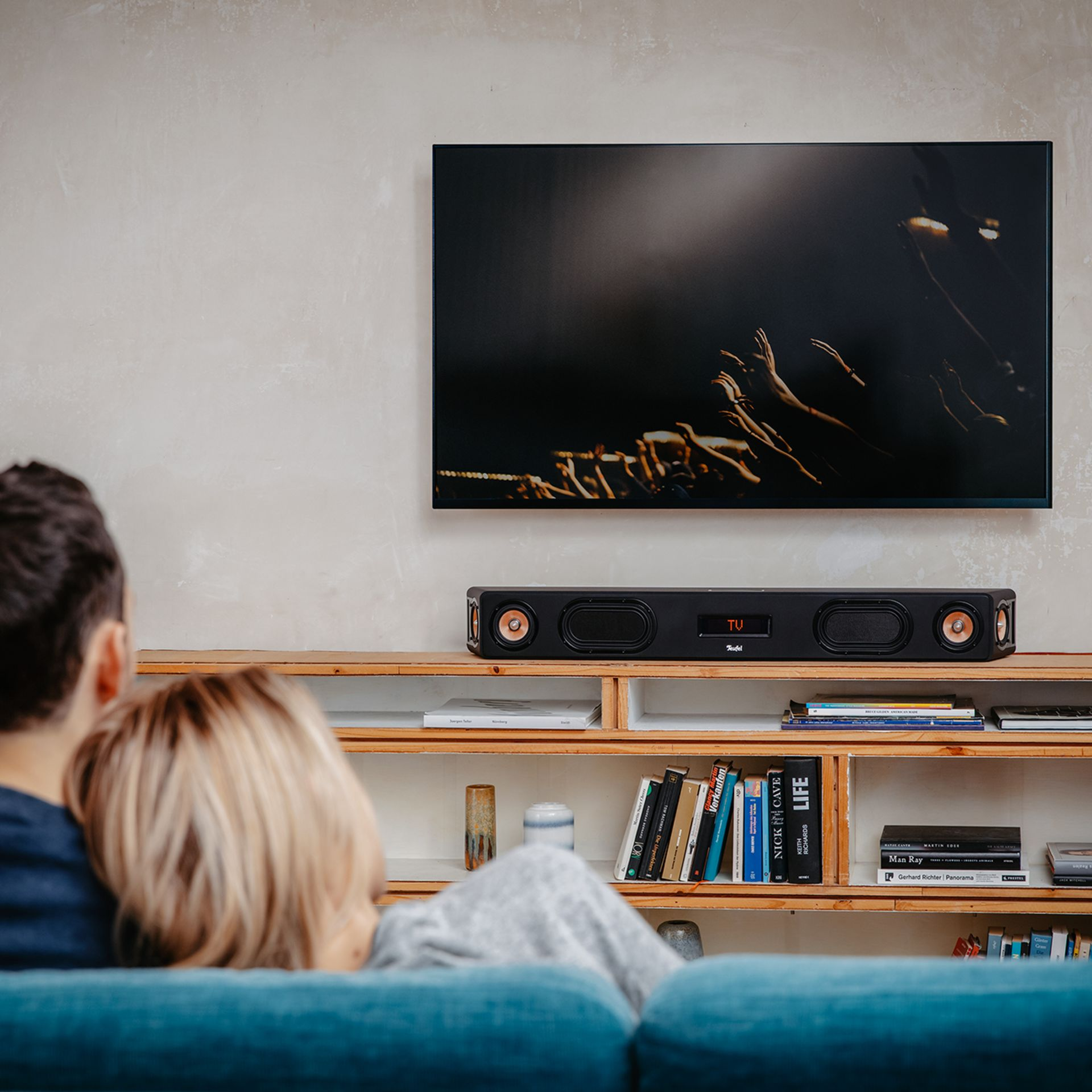 Wohnzimmer mit TV an der Wand und Soundbar. Ein Paar sitzt auf der Couch.