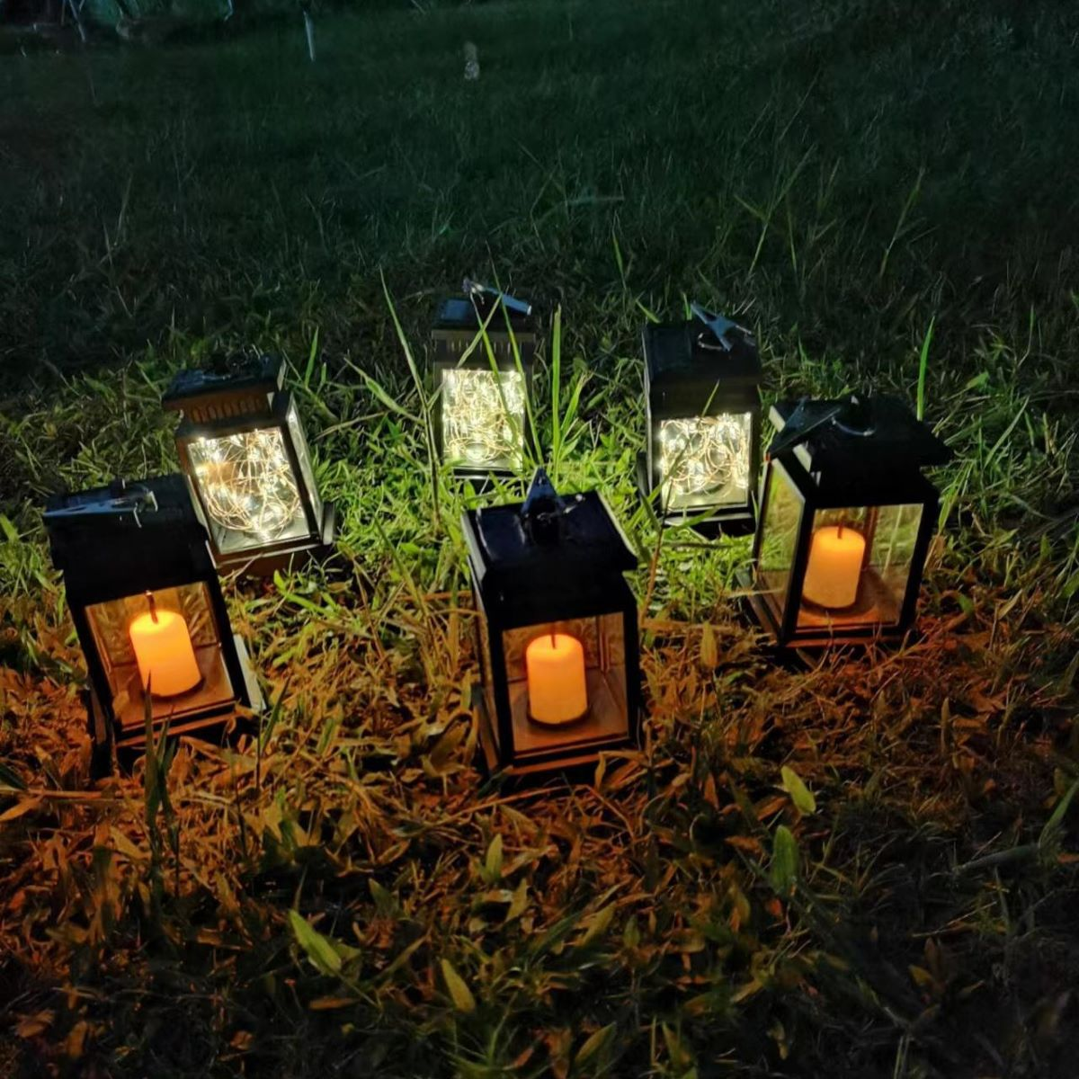 Sechs schwarze Laternen mit Kerzen im Inneren, auf Gras. Lichterketten in einigen. Im Hintergrund grünes Gras.