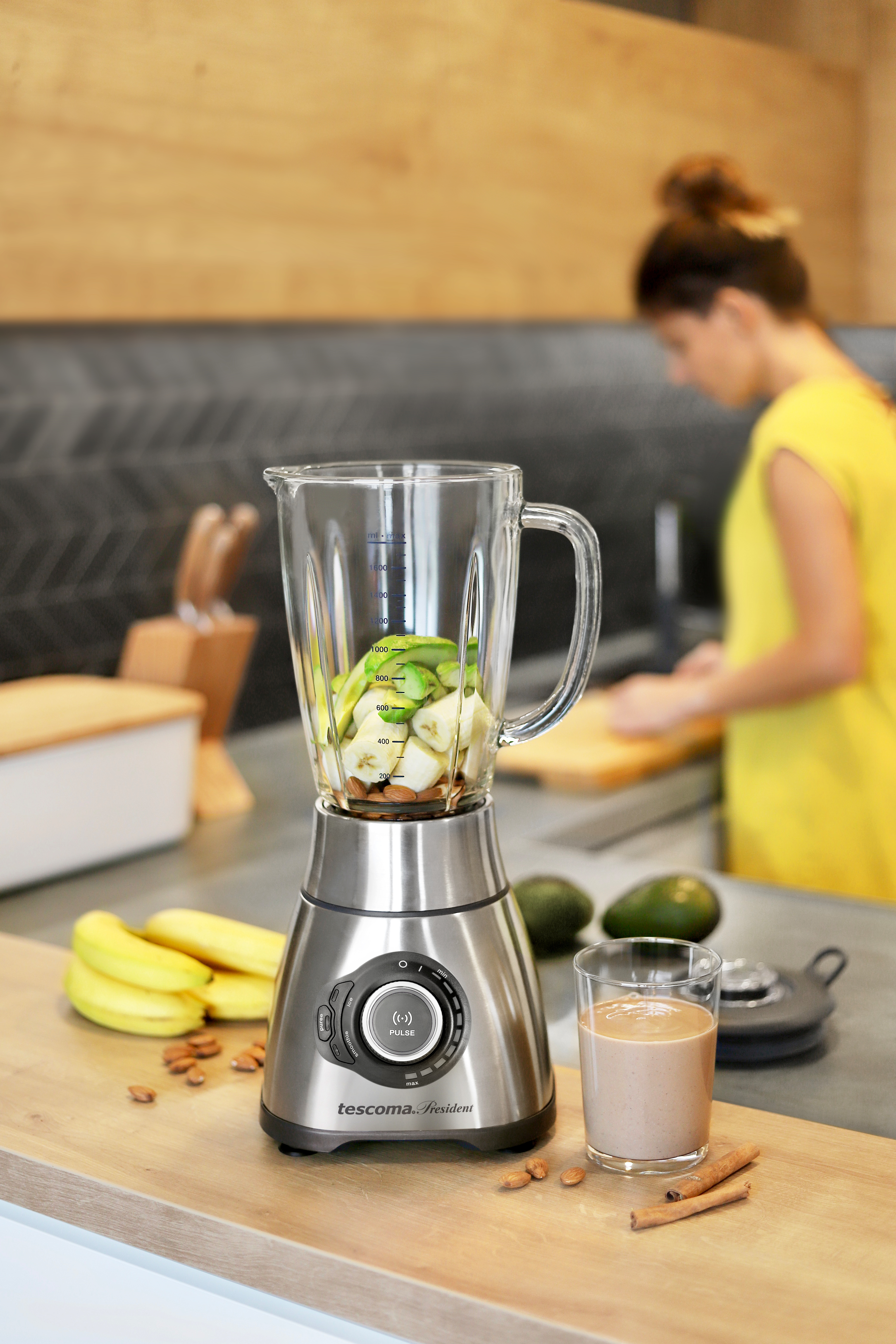 Mixer mit Früchten, Bananen, Mandeln und einem Smoothie-Glas. Frau im Hintergrund.