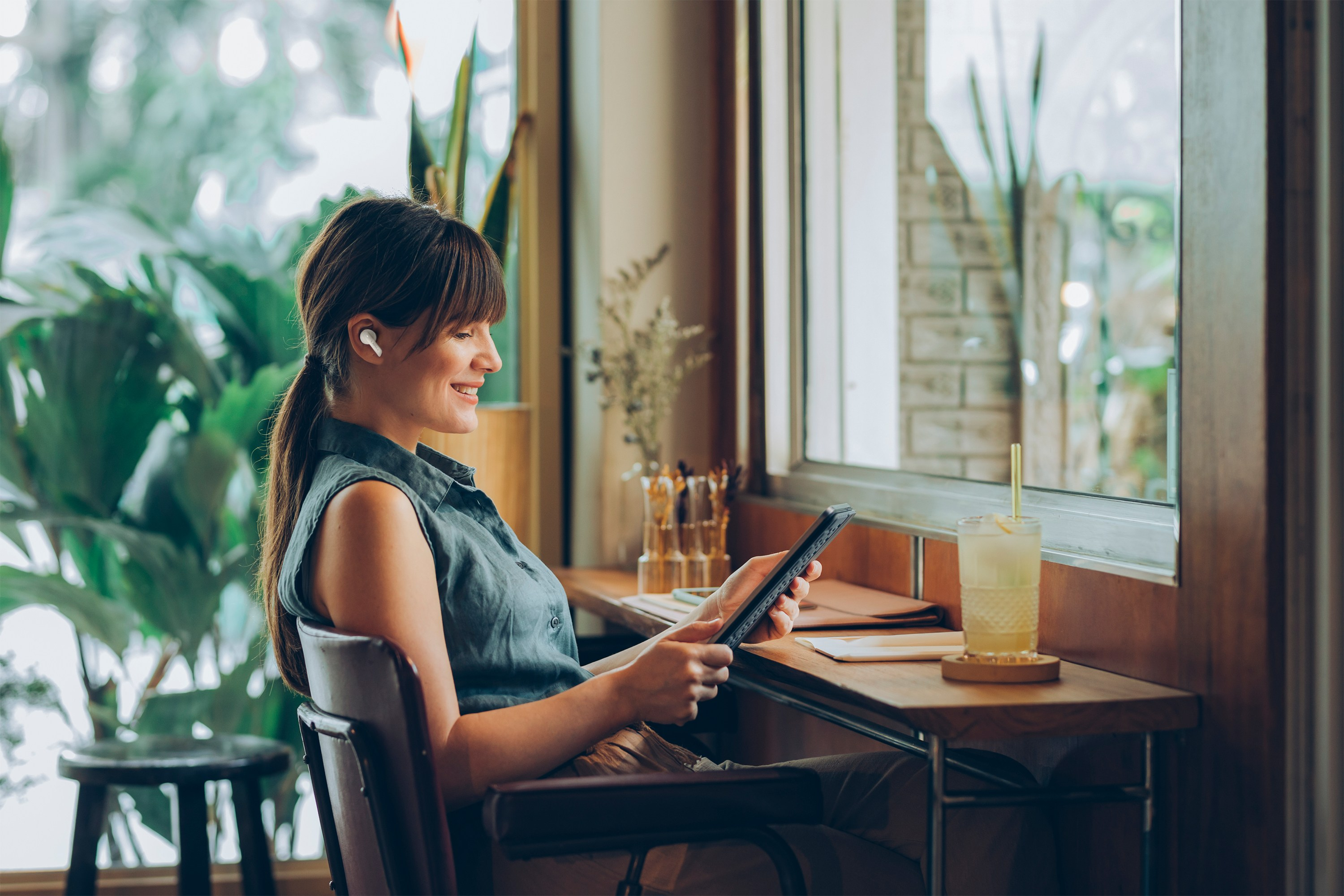 Frau im Café, mit Ohrhörern, benutzt ein Tablet, in der Nähe eines Fensters mit Getränk.