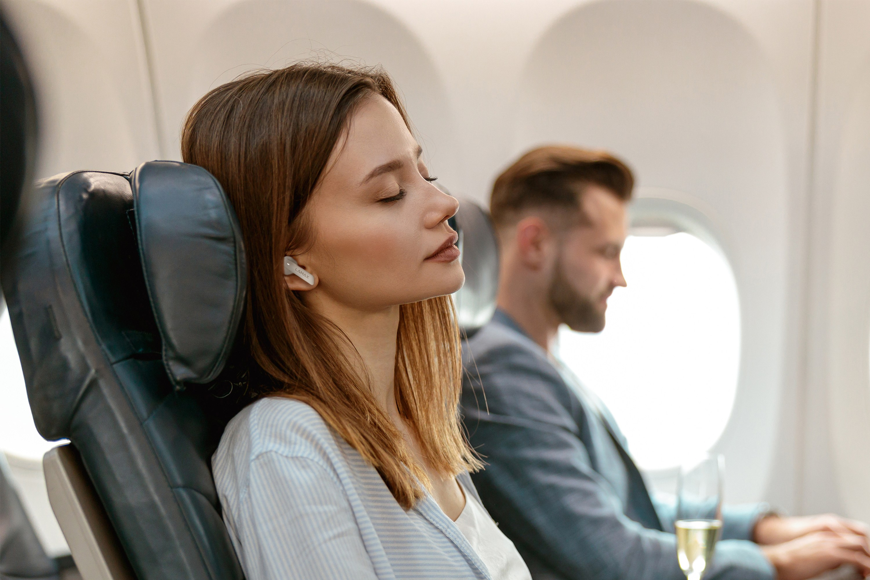 Frau und Mann in einem Flugzeug. Frau mit geschlossenen Augen, trägt Ohrhörer. Mann schaut nach vorne.