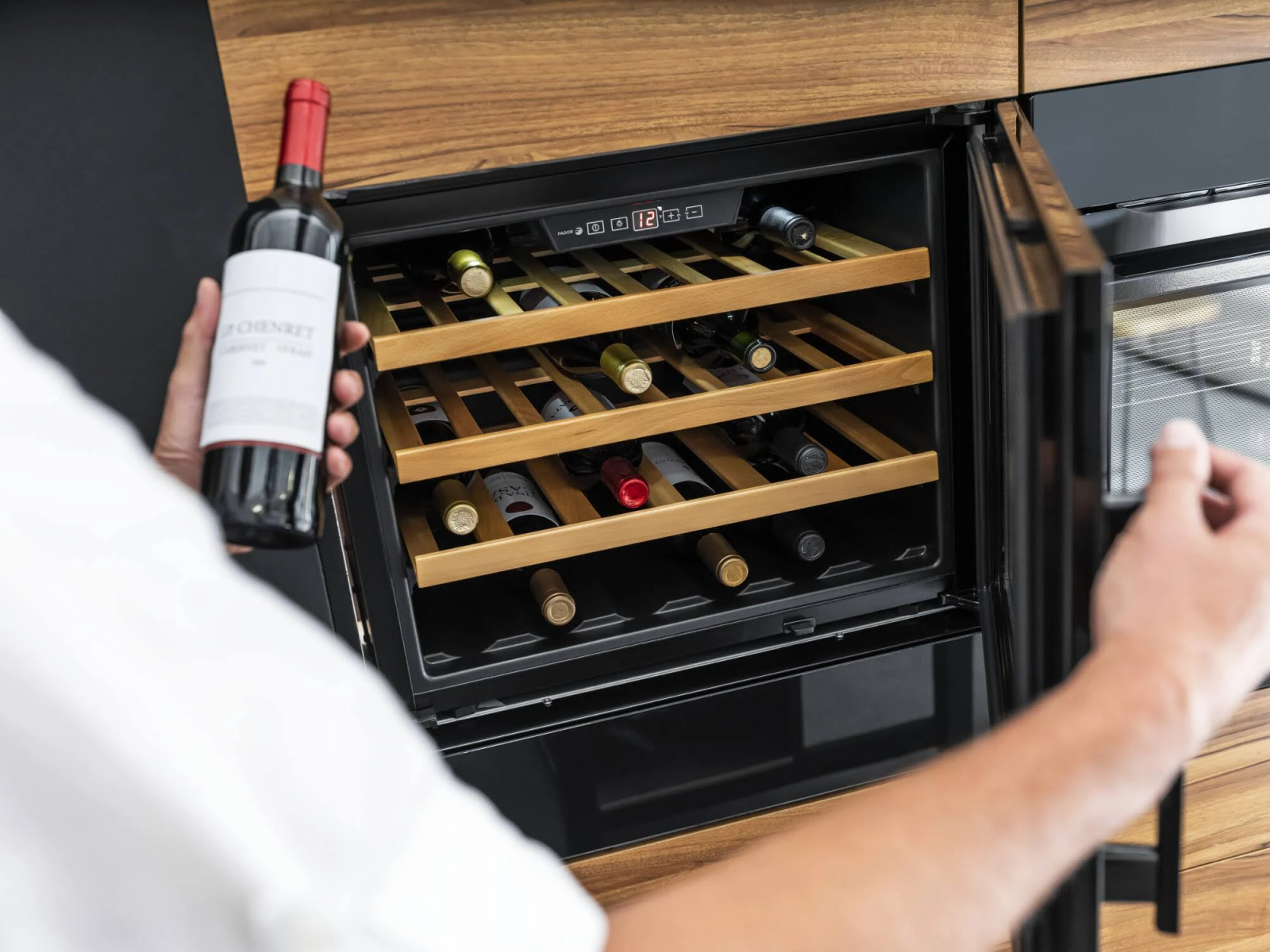 Persona che tiene una bottiglia di vino, accanto a un refrigeratore con diverse bottiglie su ripiani di legno.