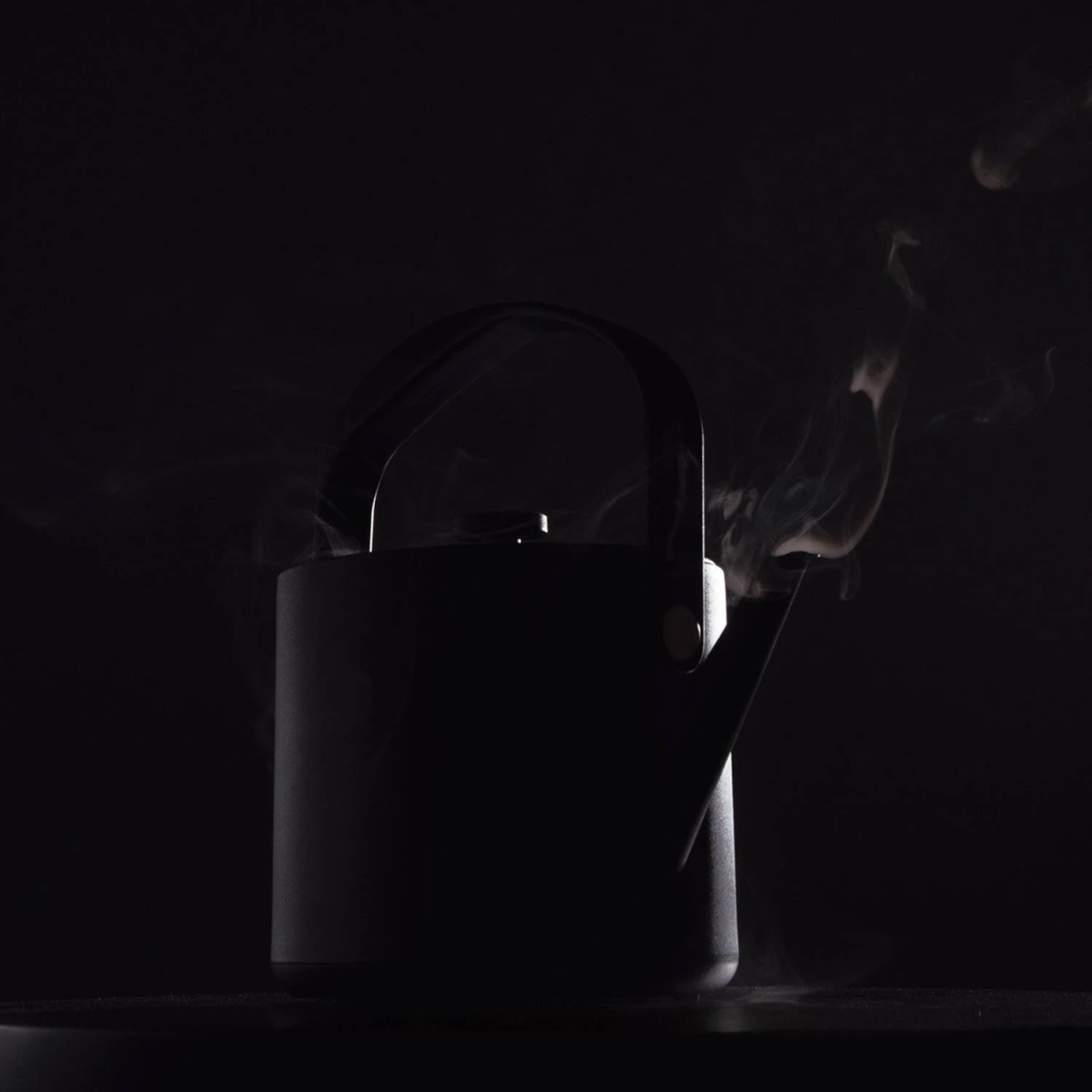 Hervidor negro con asa y boquilla, vapor saliendo de la boquilla, en un entorno oscuro.