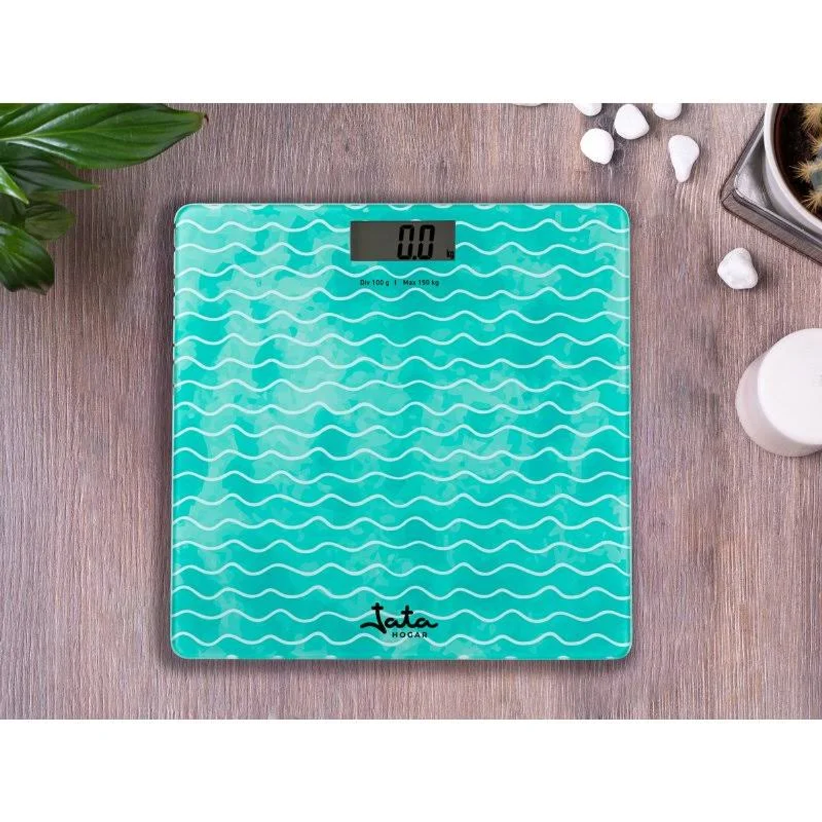 Una báscula digital turquesa con un patrón ondulado, una planta y guijarros blancos.