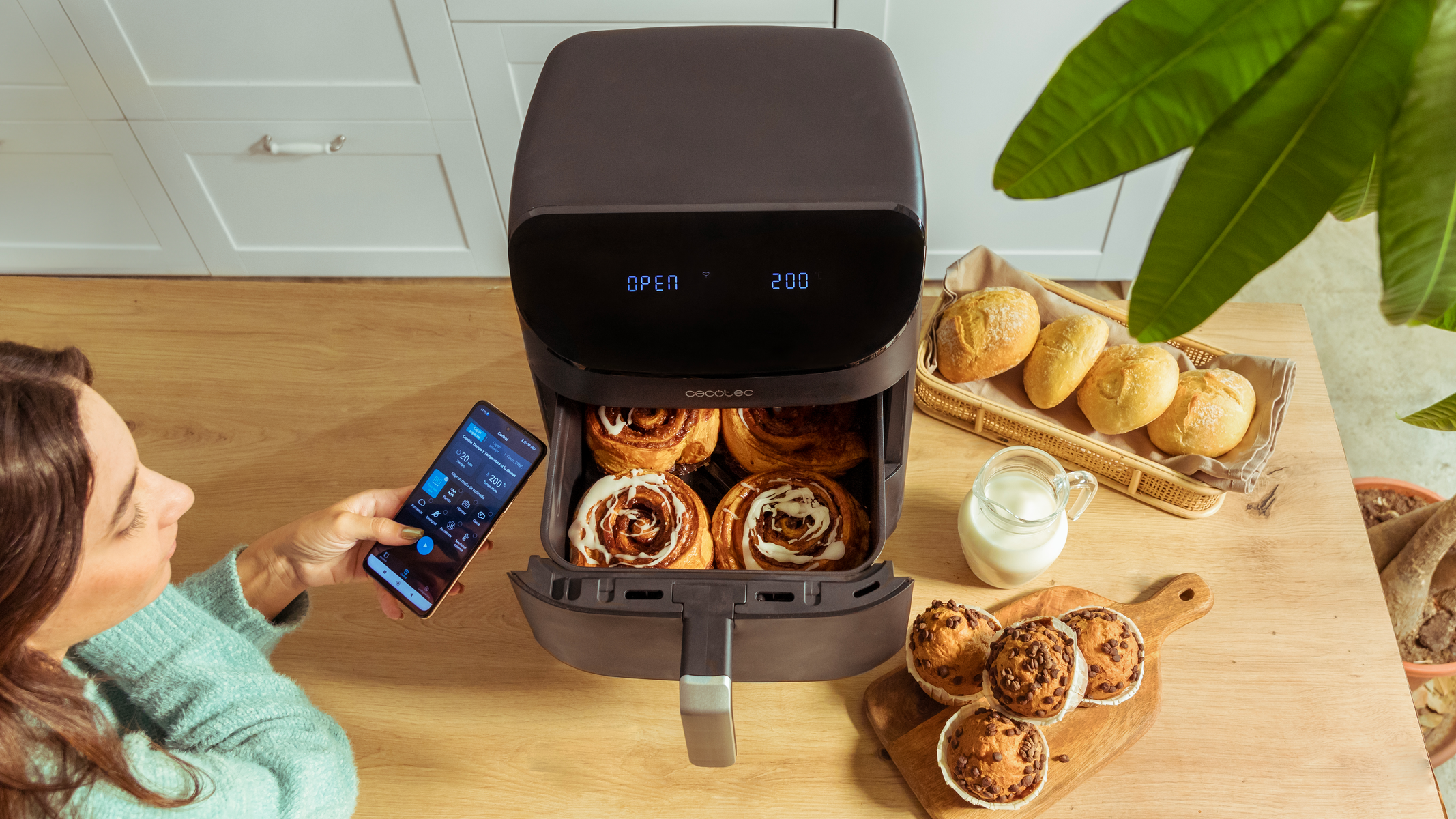 Vrouw bestuurt airfryer met smartphone, met brood en muffins.