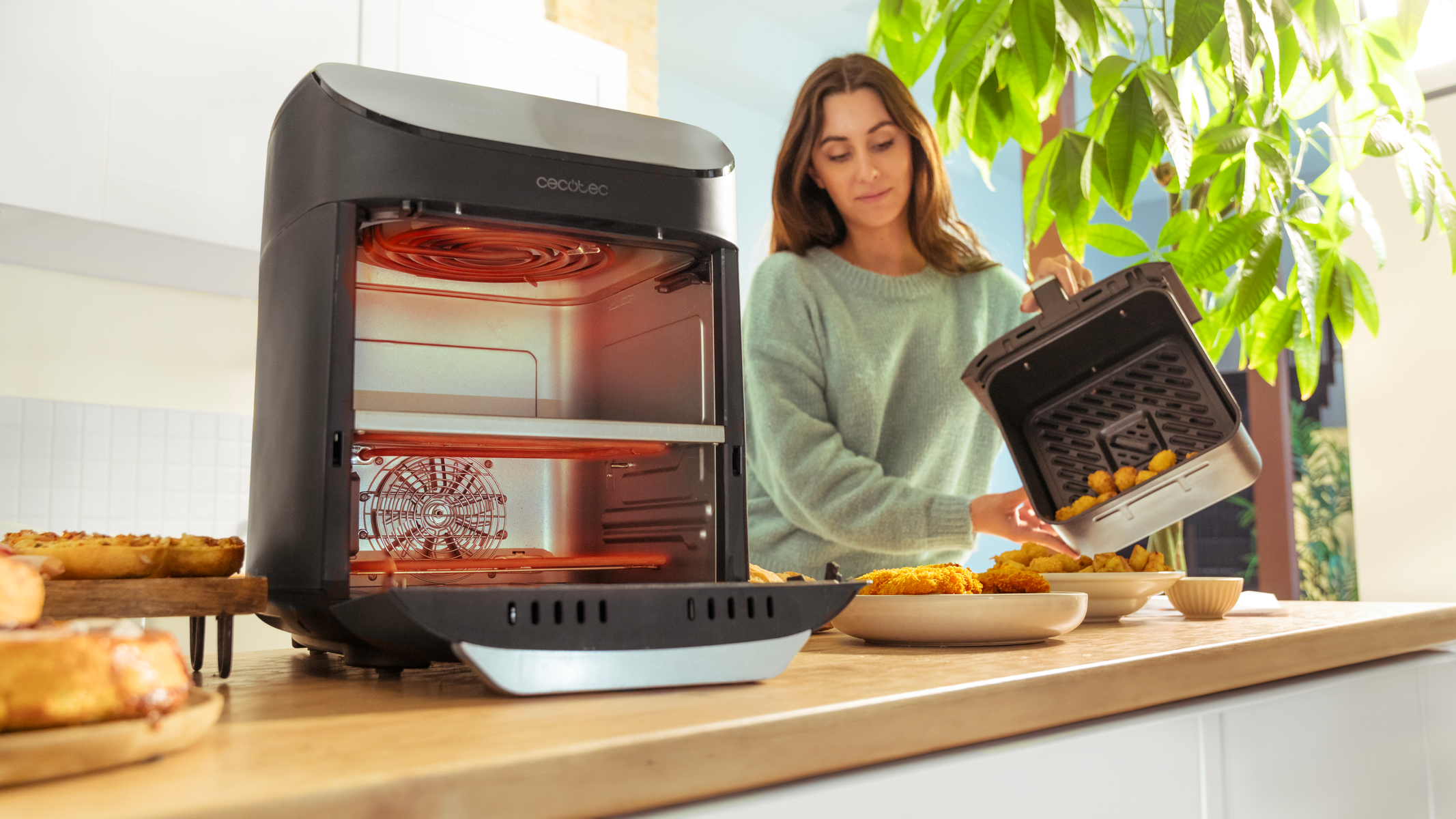 Vrouw gebruikt een airfryer. Gebakken eten op borden, oven links. Groene planten op de achtergrond.