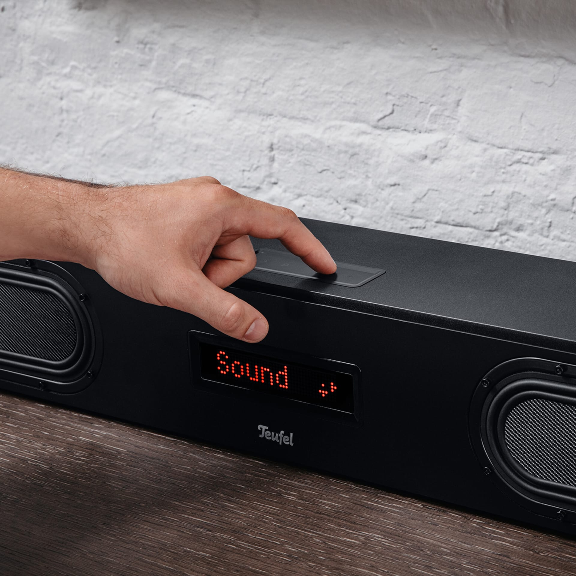 Schwarze Soundbar mit roter Anzeige. Hand drückt eine Taste. Holztisch und Ziegelmauer.
