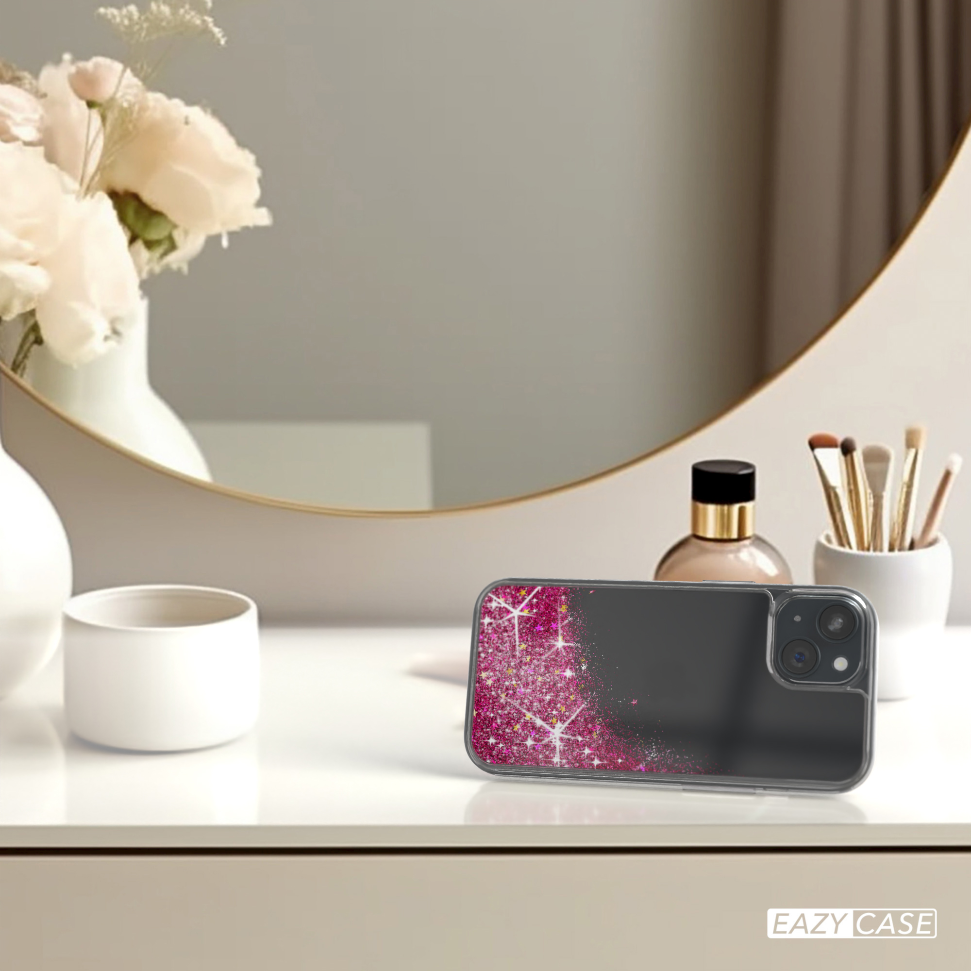 Eine Handyhülle mit rosa Glitzer auf einem Tisch mit Blumen, einem Spiegel und Schönheitsprodukten.
