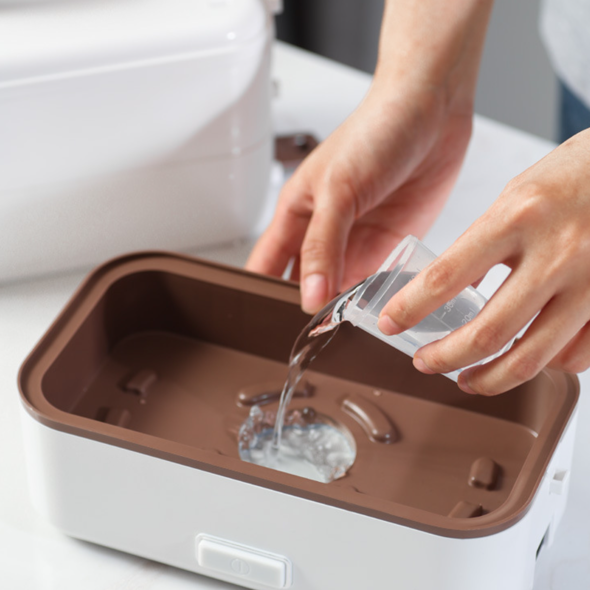Hände gießen Wasser aus einem Messbecher in eine weiße und braune Lunchbox.