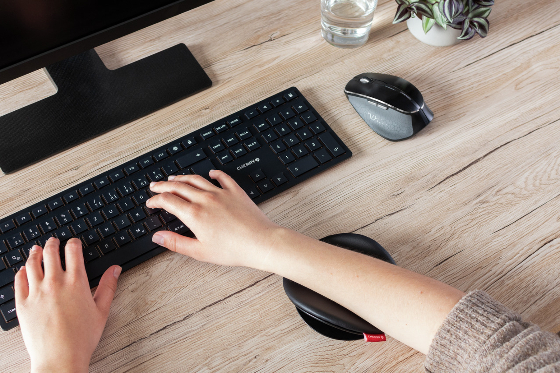 Eine Person, die einen Computer mit einer schwarzen Handgelenkstütze mit einem roten Cherry-Logo-Etikett auf einem Schreibtisch aus Holz benutzt.