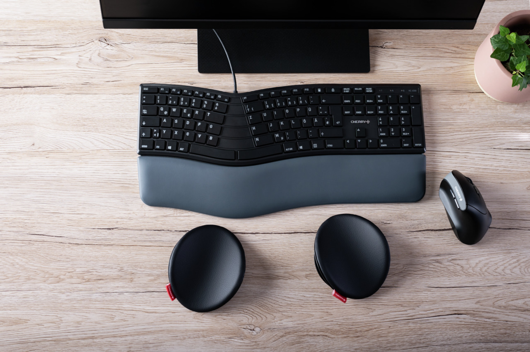 Eine geschwungene Tastatur, Maus und Handgelenkstützen auf einem Holztisch. Ein Monitor und eine Pflanze sind ebenfalls vorhanden.
