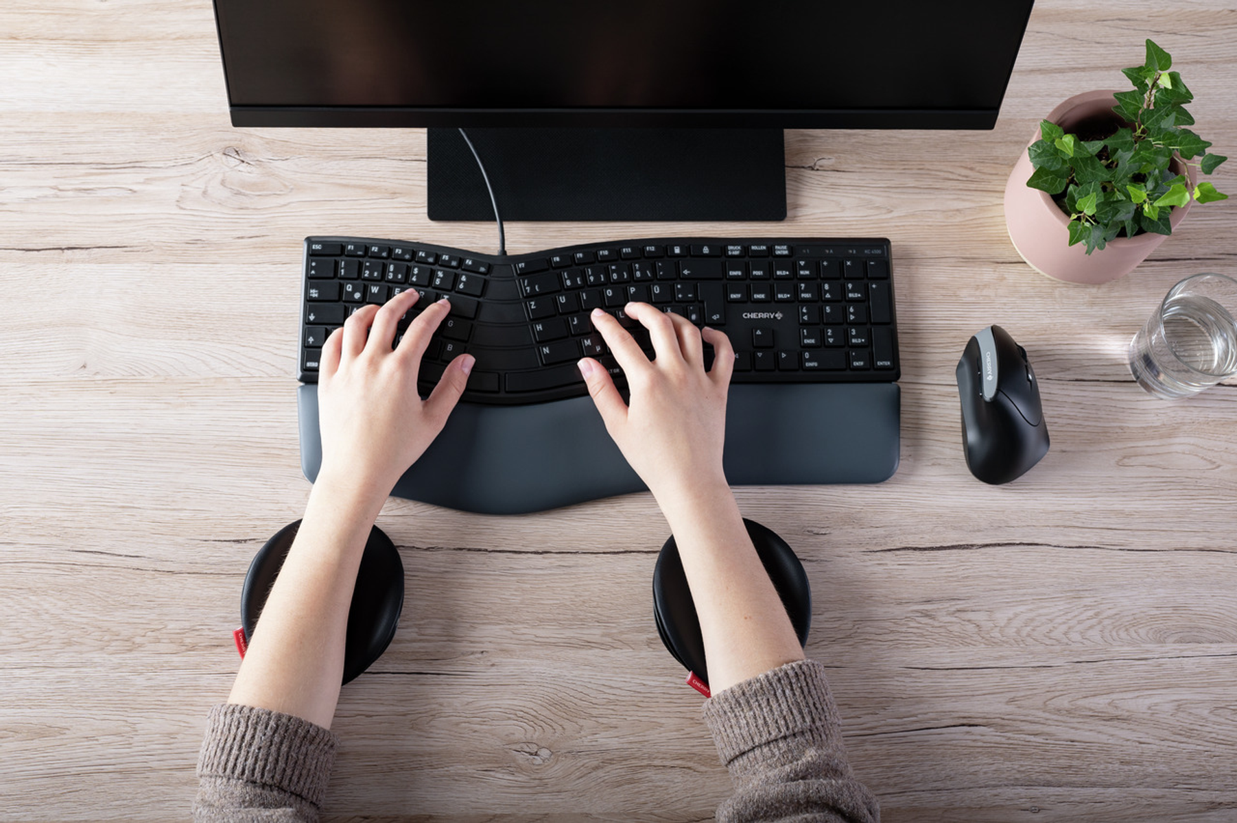 Hände tippen auf einer geschwungenen Tastatur. Maus und Handgelenkstützen befinden sich auf einem Holztisch, mit Monitor und Pflanze.