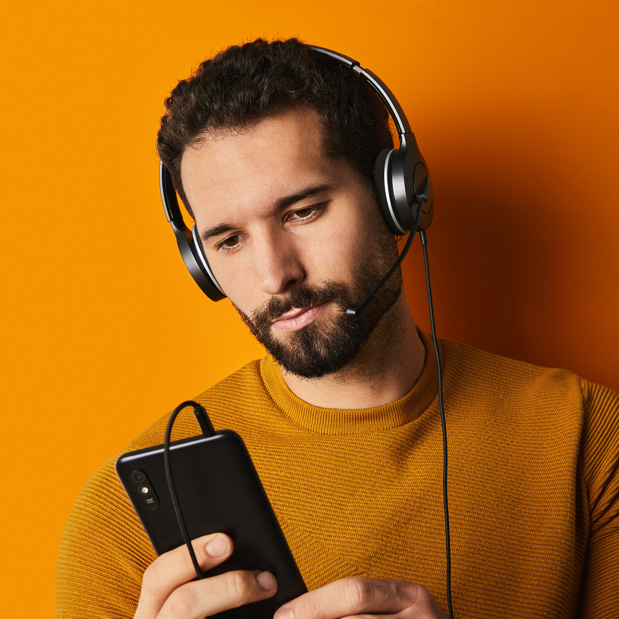 Un hombre con barba que lleva auriculares y mira un smartphone. Está sobre un fondo naranja.