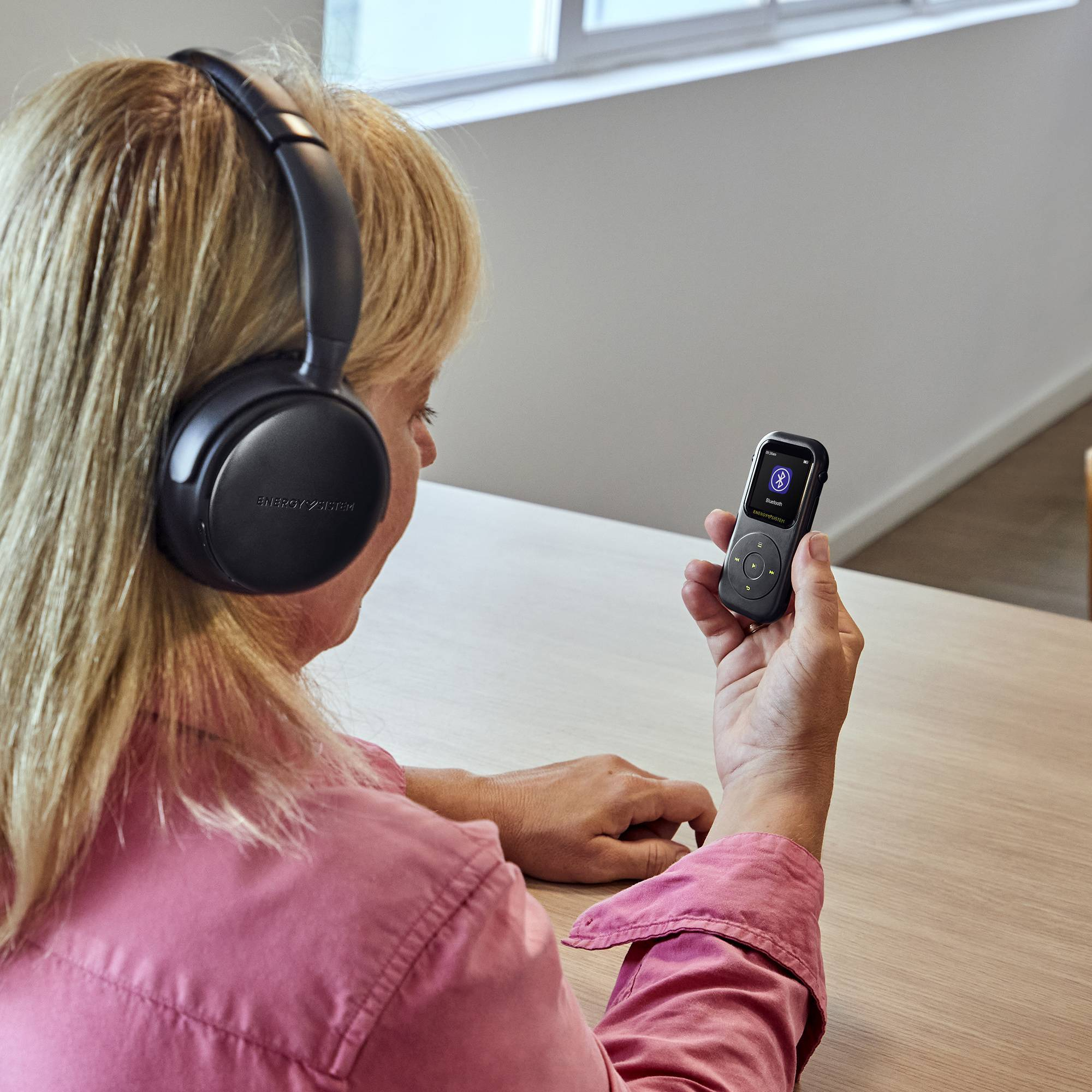 Una mujer usa auriculares negros mientras sostiene un pequeño dispositivo negro. La pantalla del dispositivo es visible.
