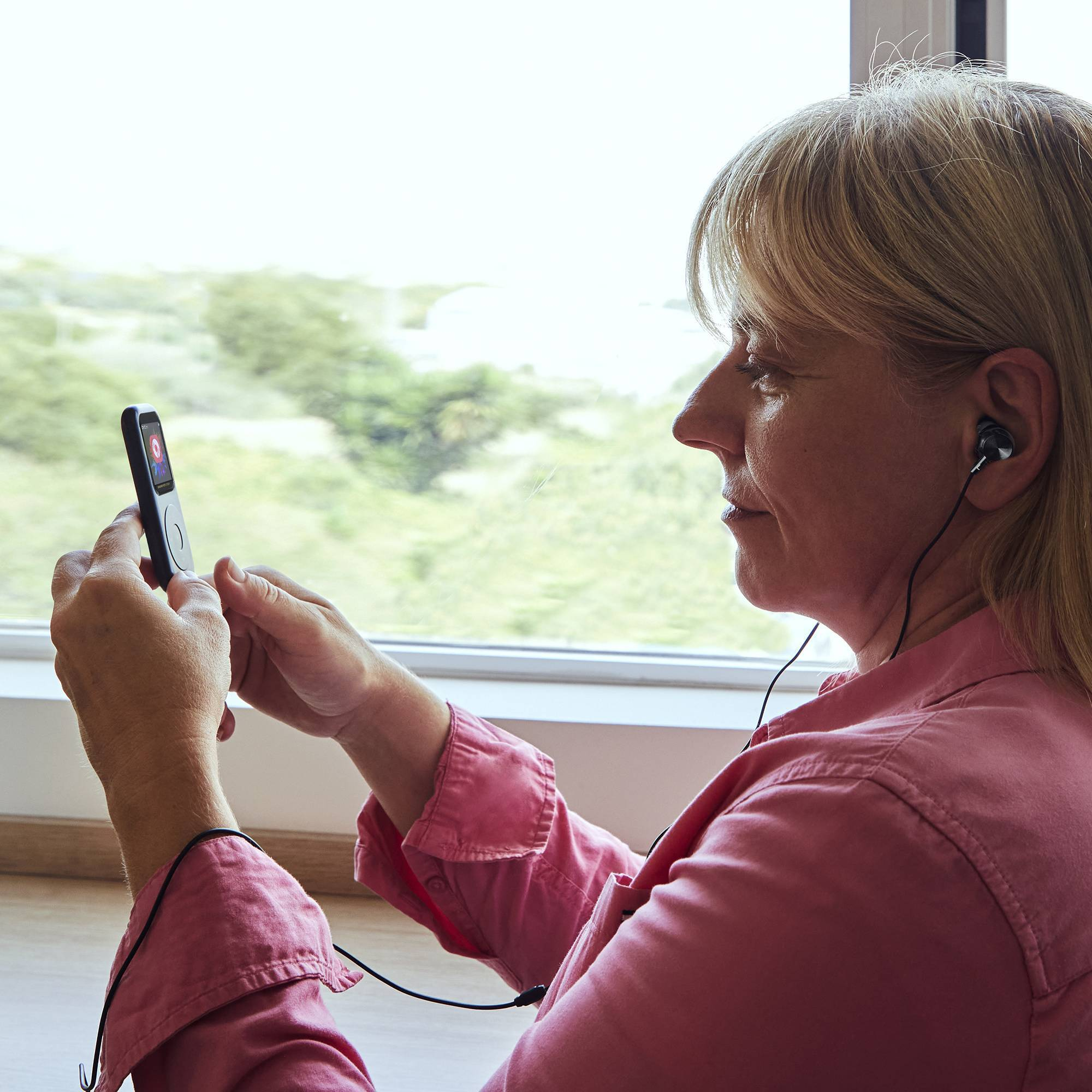 Una mujer escuchando música en un pequeño reproductor, con auriculares, cerca de una ventana.