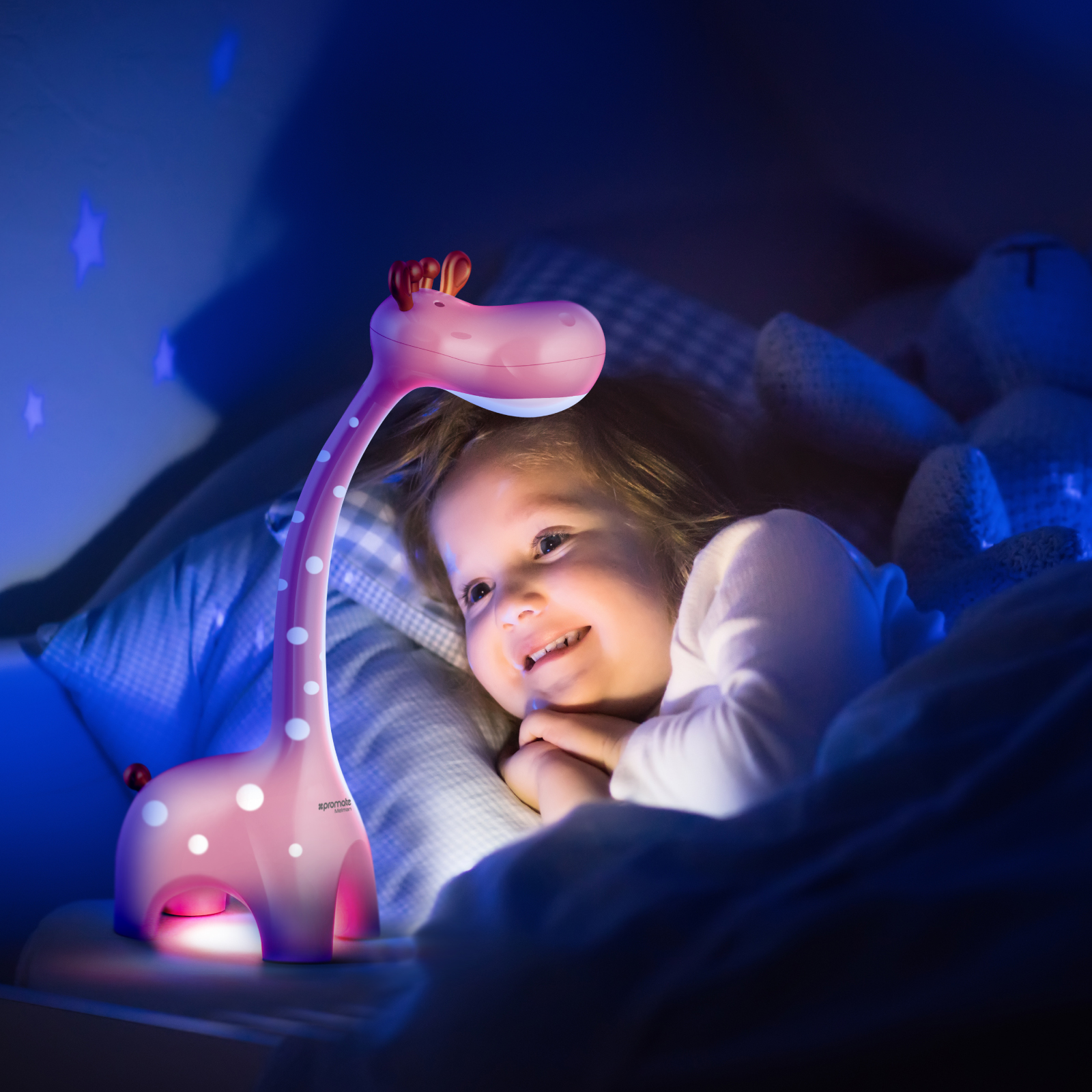 Una lámpara de jirafa rosa ilumina a un niño acostado en la cama. El niño sonríe, rodeado de almohadas y una manta.