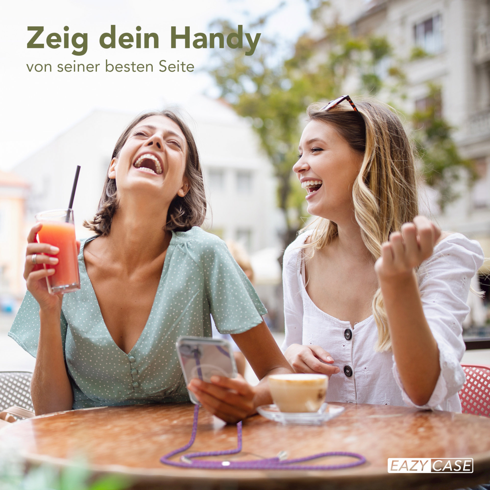 Zwei lachende Frauen an einem Tisch, halten Getränke und ein Telefon. Sie befinden sich im Freien.
