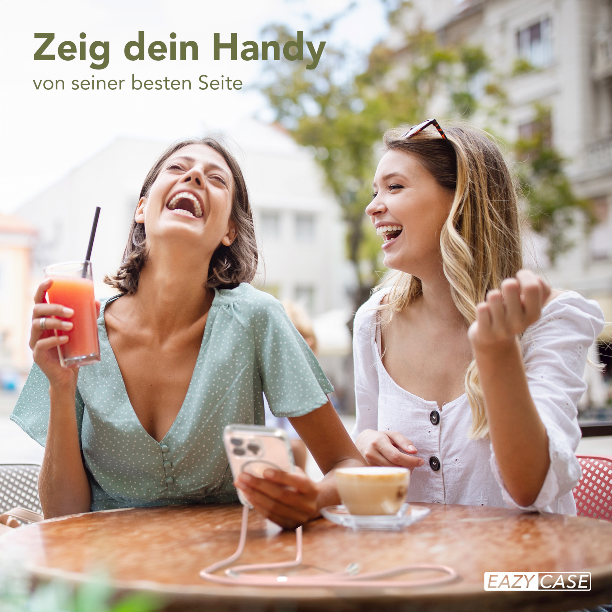 Zwei Frauen lachen in einem Straßencafé, eine hält ein Handy mit Hülle, Getränke auf dem Tisch.