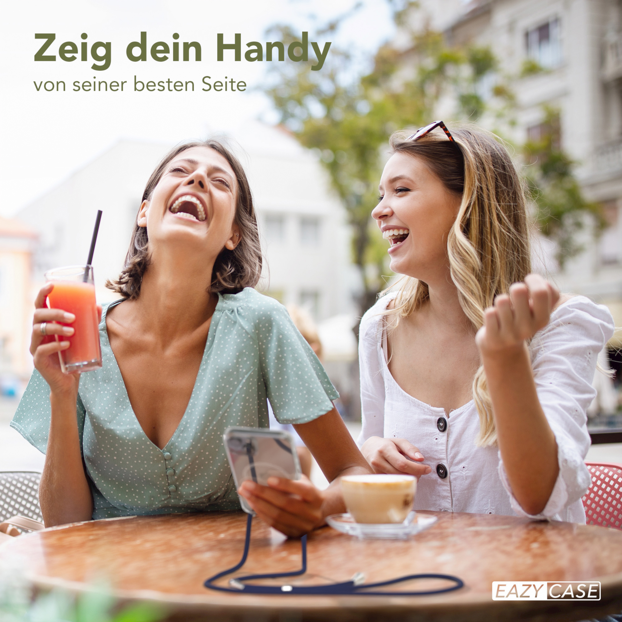 Zwei lachende Frauen, eine hält ein Telefon mit einem Riemen, in einem Café, mit Getränken und einem Tisch.